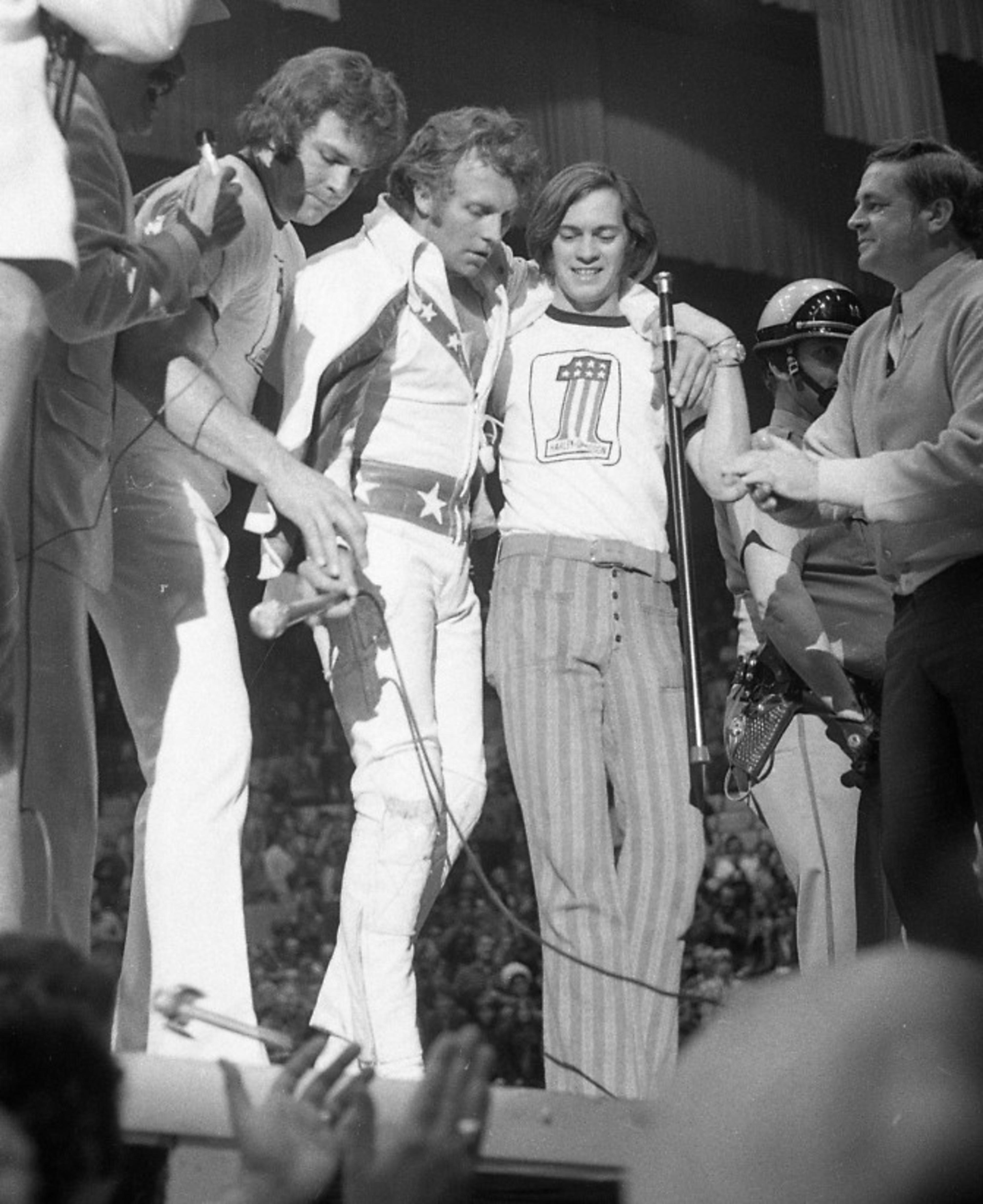 A black and white photo of a group on stage, with one person in a star-patterned outfit in the center, surrounded by others and a crowd below.