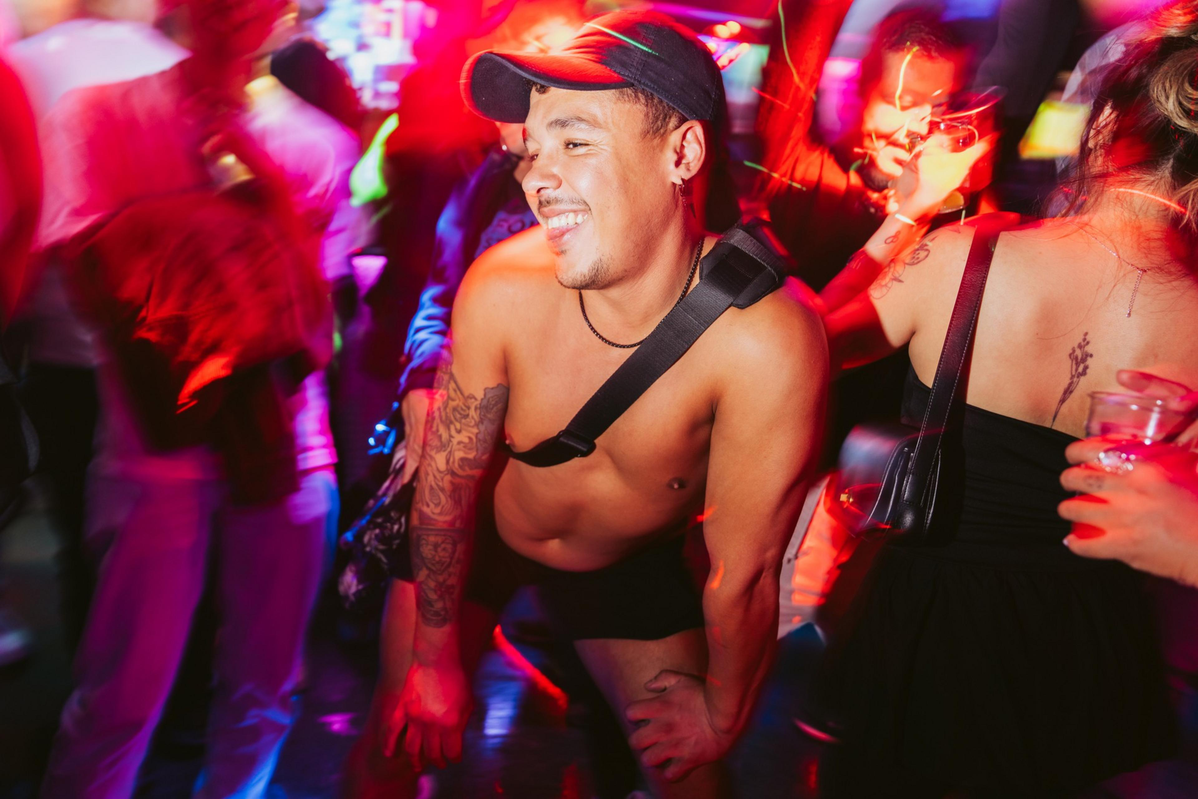 A shirtless man with a tattoo dances energetically in a lively, colorful nightclub, surrounded by other party-goers. The lighting is vivid and dynamic.