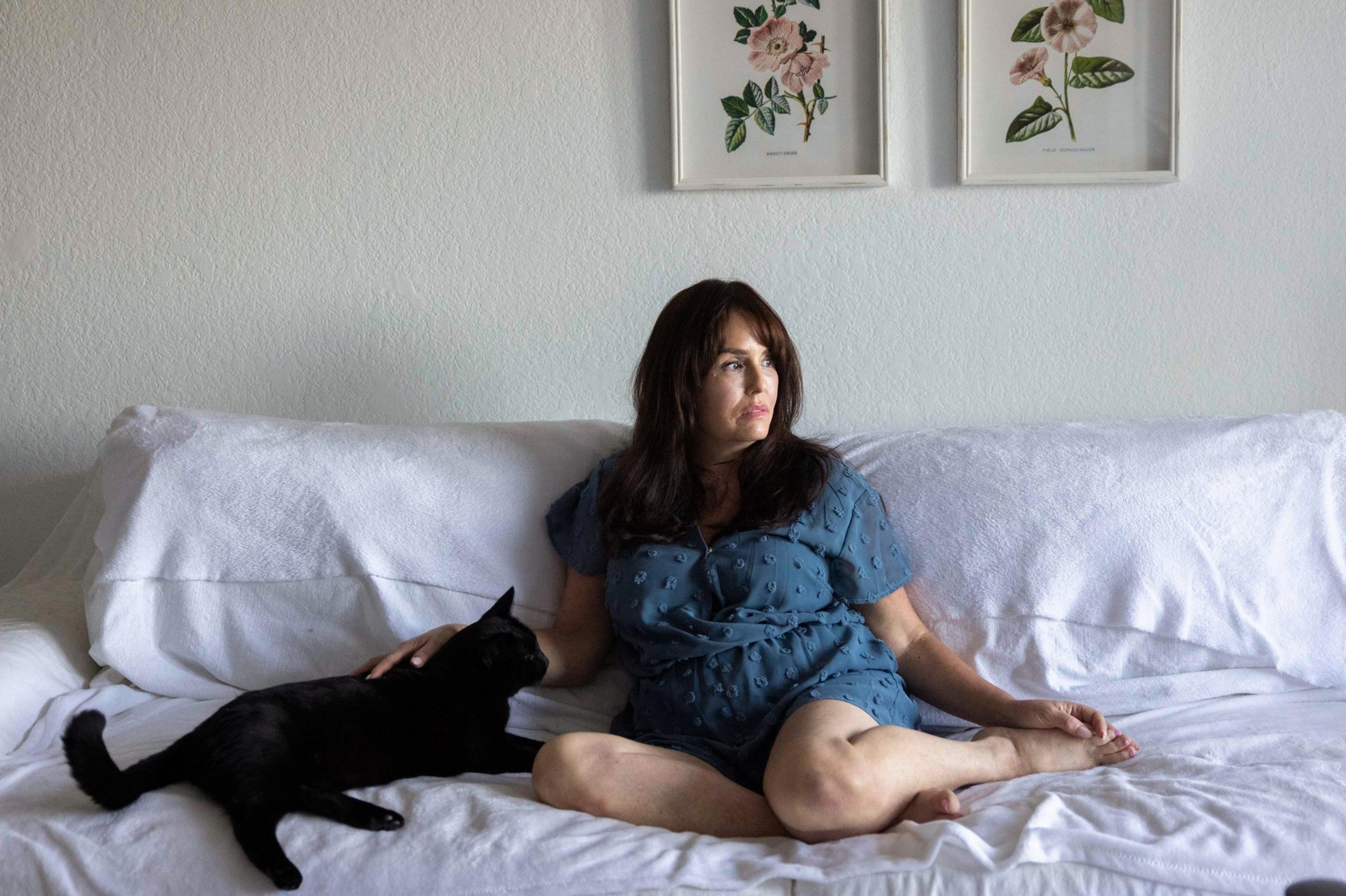 A woman in a blue dress sits on a white couch, petting a black cat. She looks to the side. Two floral prints hang on the wall above her.