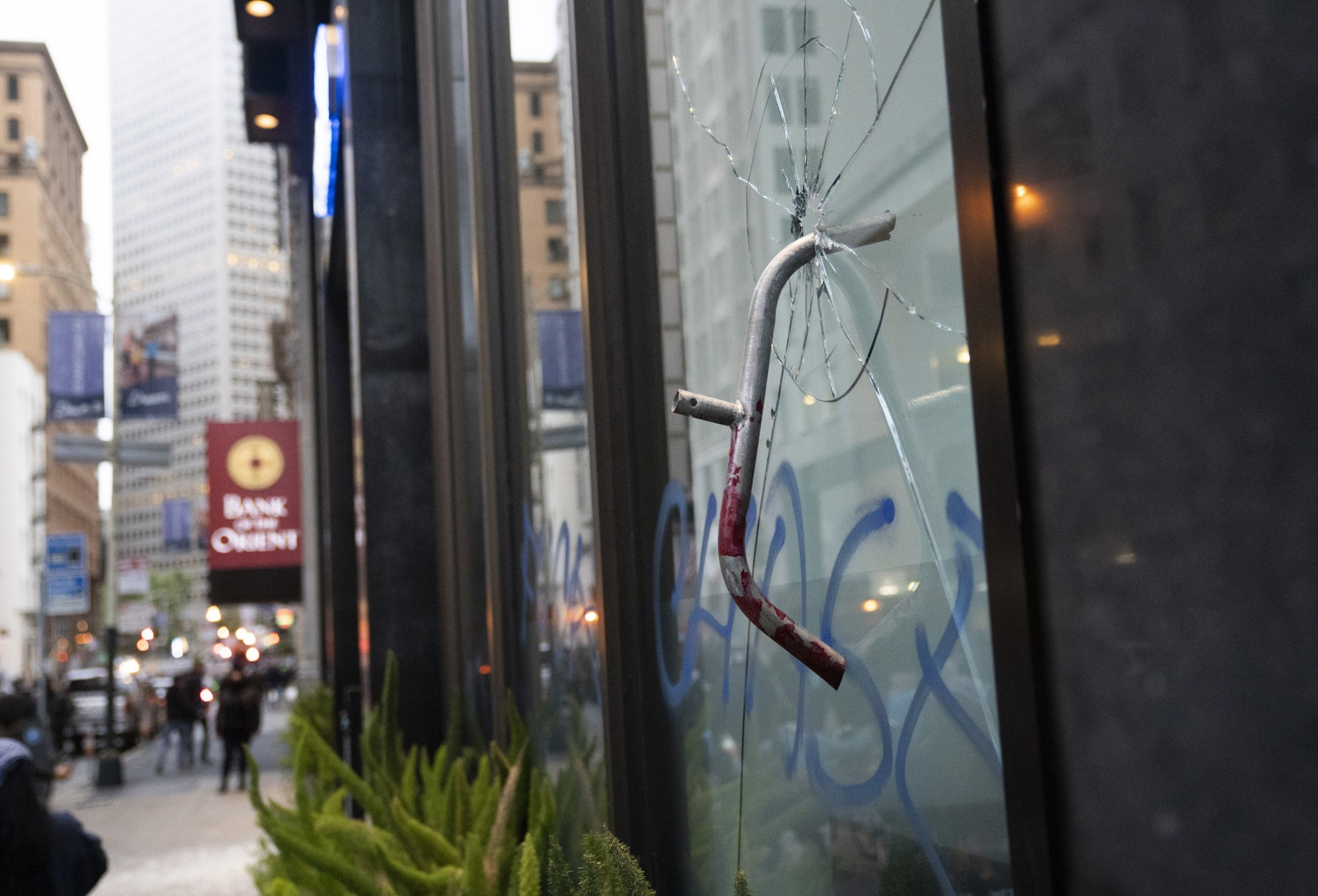A cracked window with a metal rod protruding from it is seen. The background depicts a city street with pedestrians and various buildings.