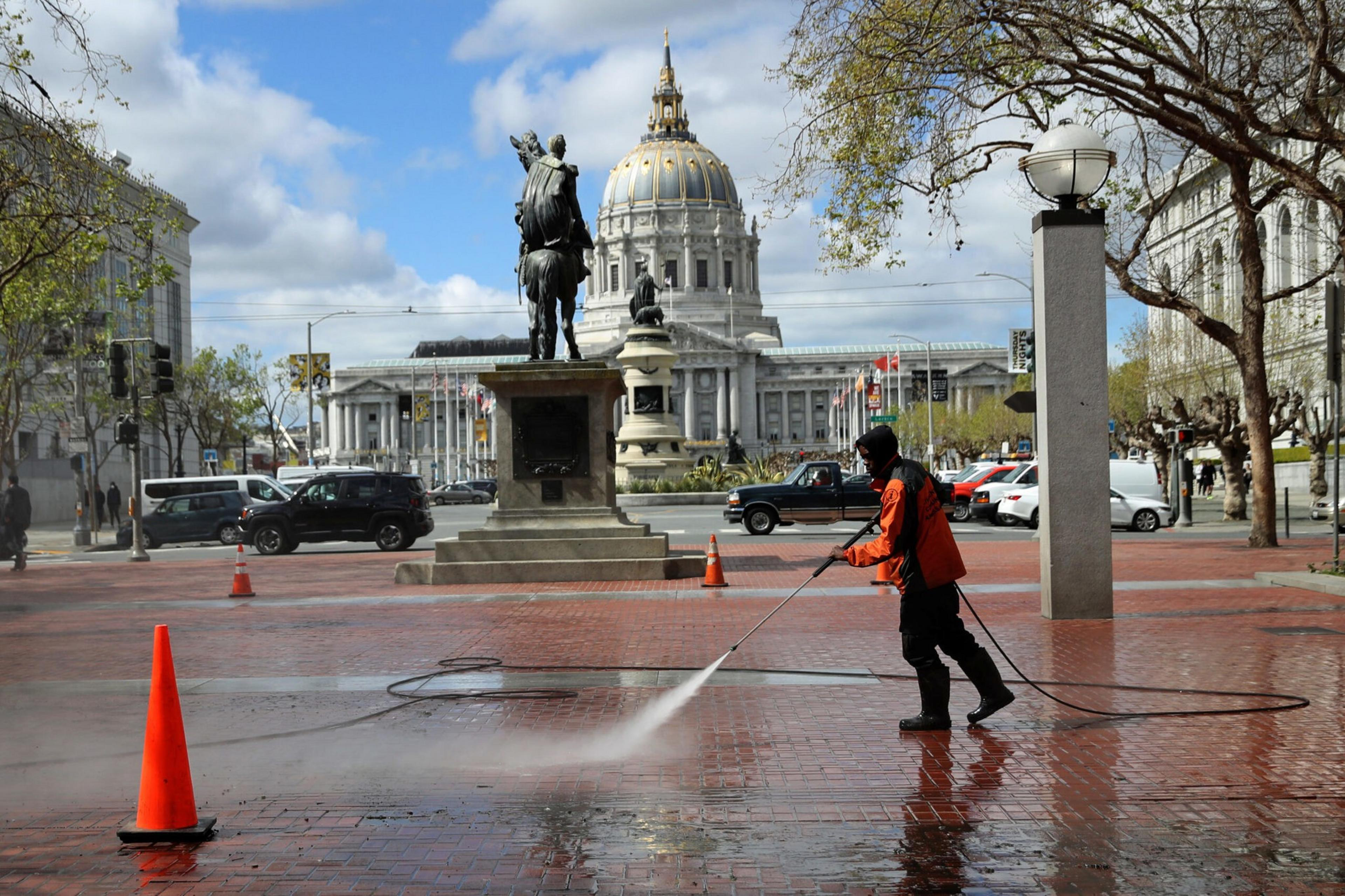 A worker in an orange jacket power washes a red brick plaza near a statue, with a grand domed building and cars in the background under a partly cloudy sky.