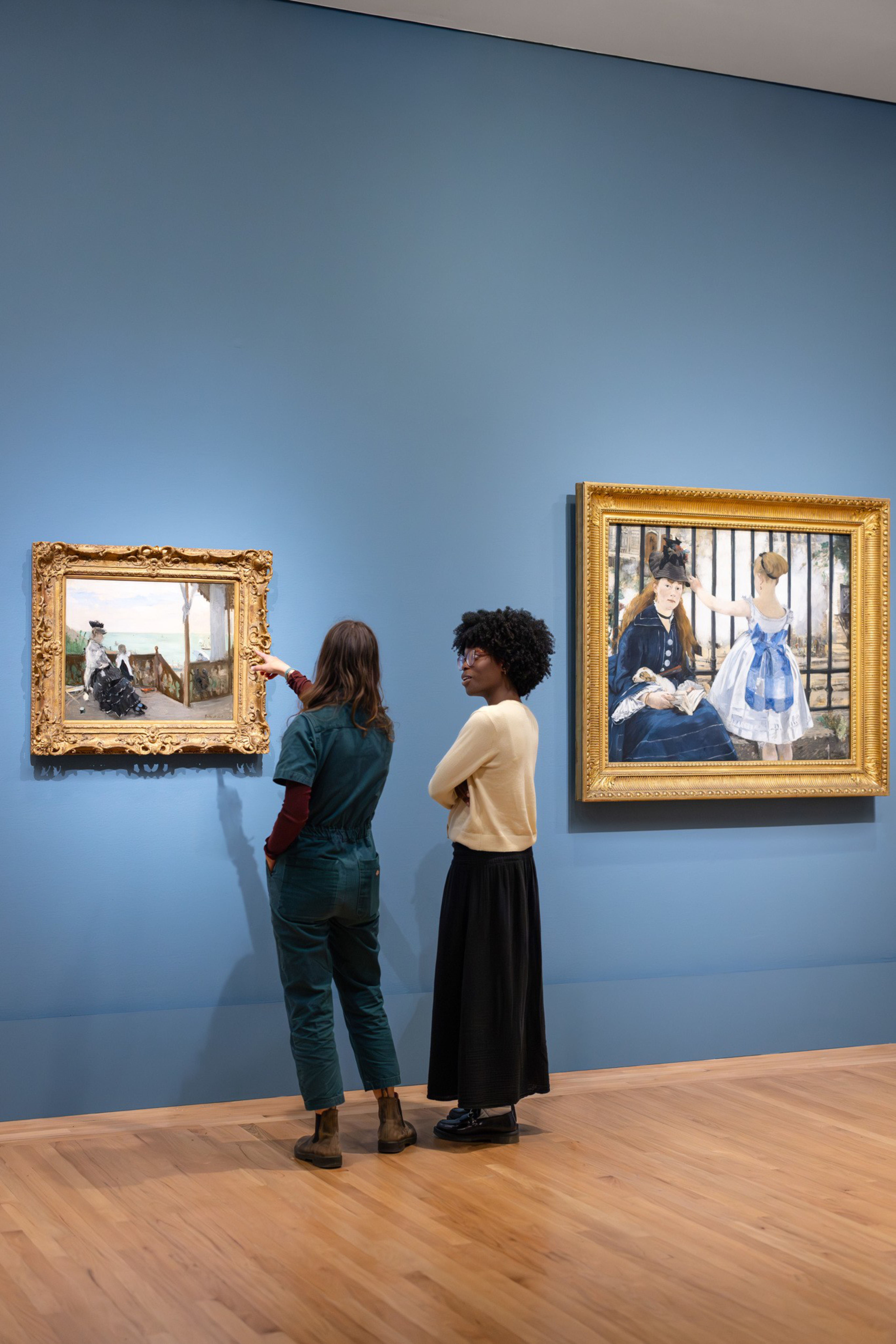 Two women stand in an art gallery with blue walls, observing and discussing two framed paintings featuring people in historical attire.