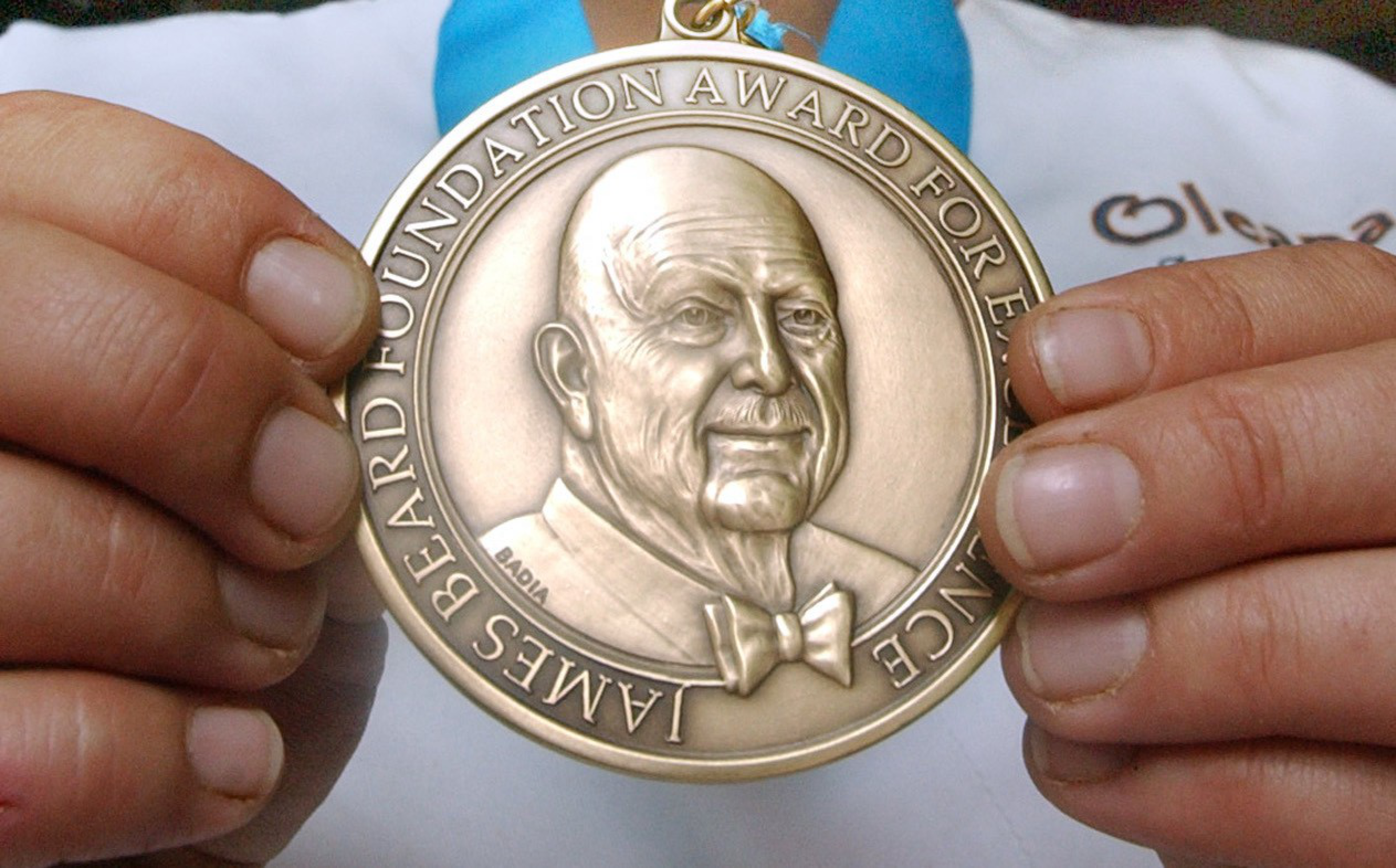 A bronze medal with an engraved portrait of a man wearing a bow tie, held by two hands, reads "James Beard Foundation Award for Excellence."