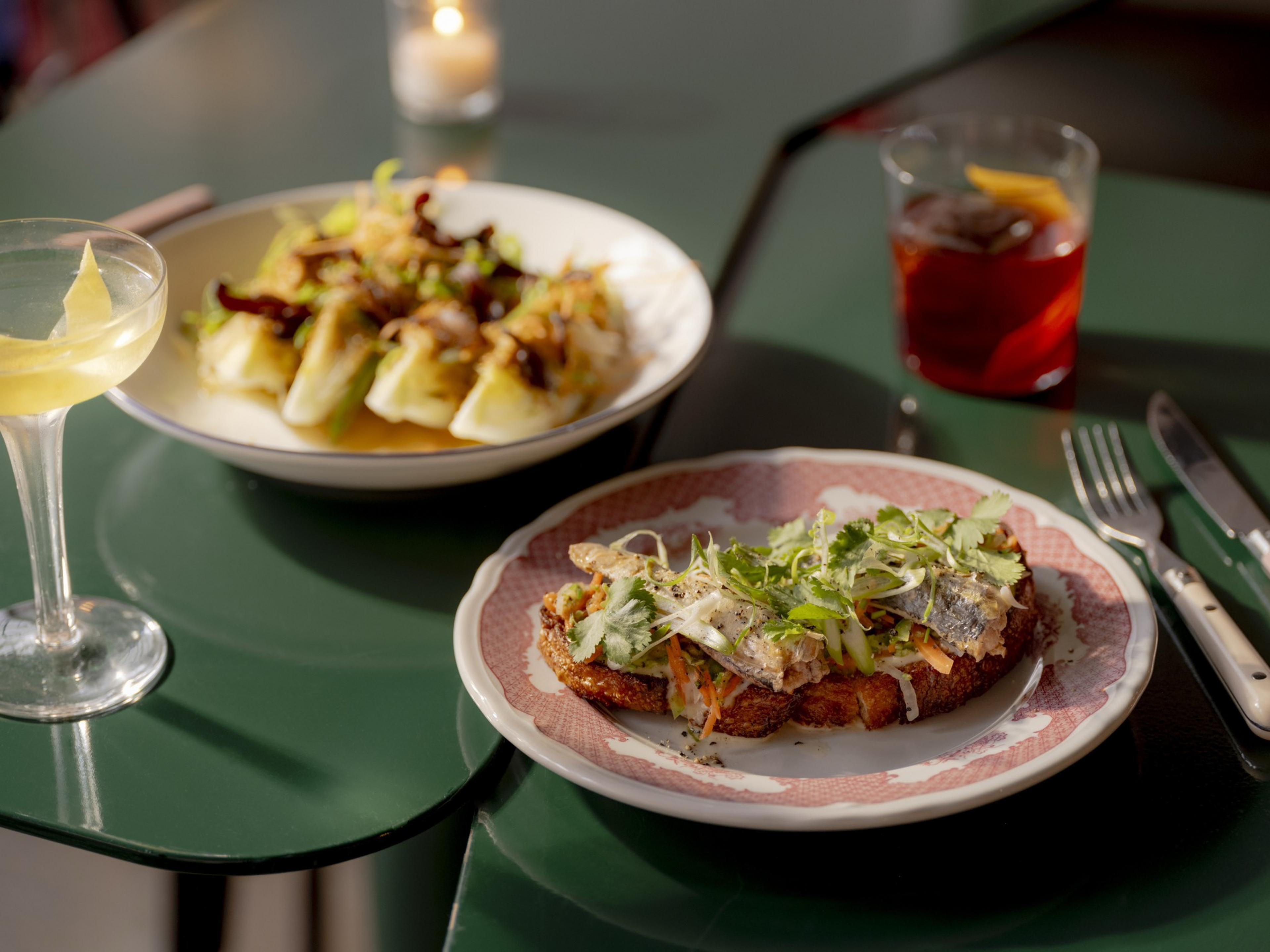 A green table holds a plate of toast with fish and greens, another with stuffed vegetables, a cocktail with a lemon twist, and a red drink. Candle ambiance is present.