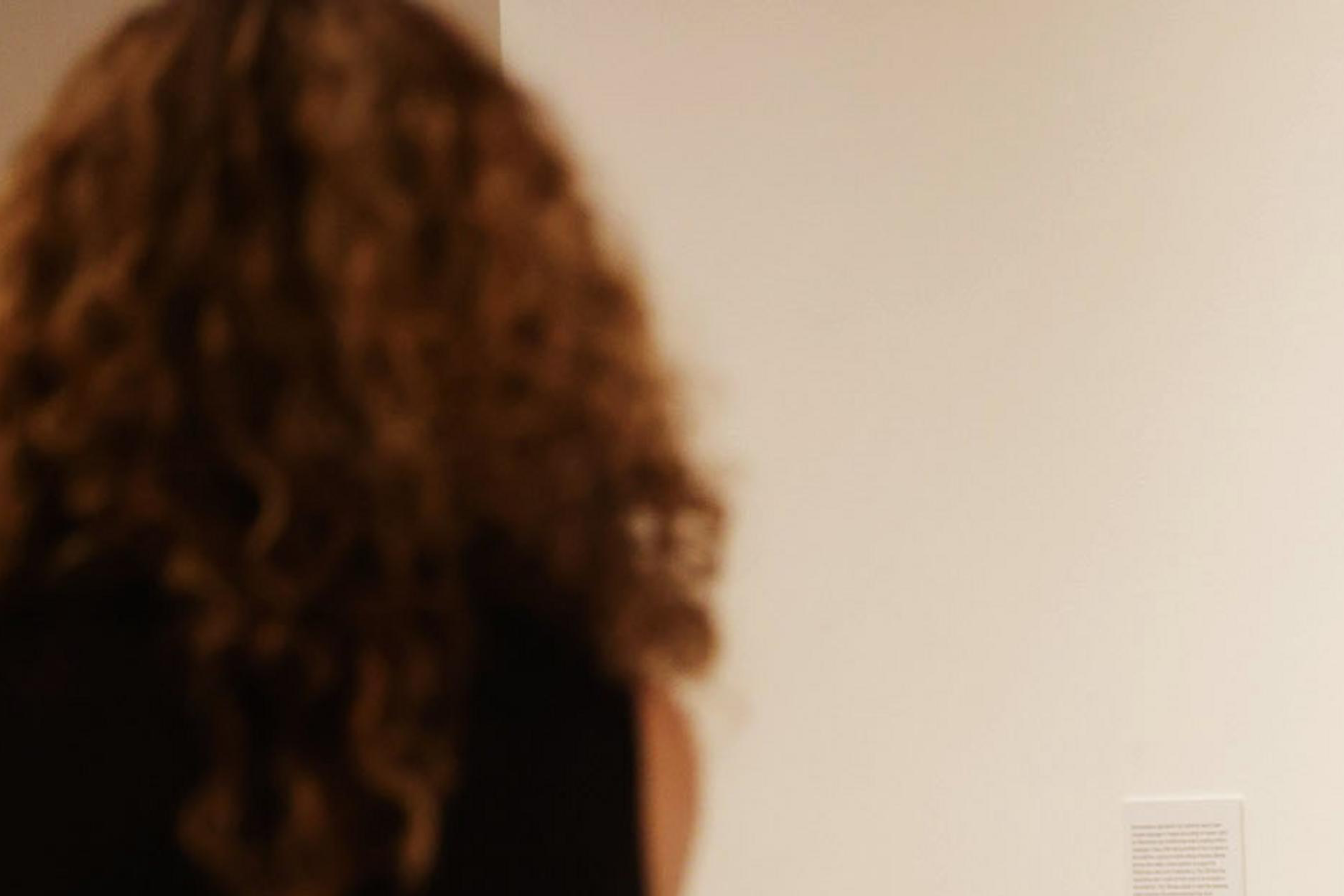 A person with curly hair, dressed in a sleeveless top, stands in front of a beige wall reading two small text panels.