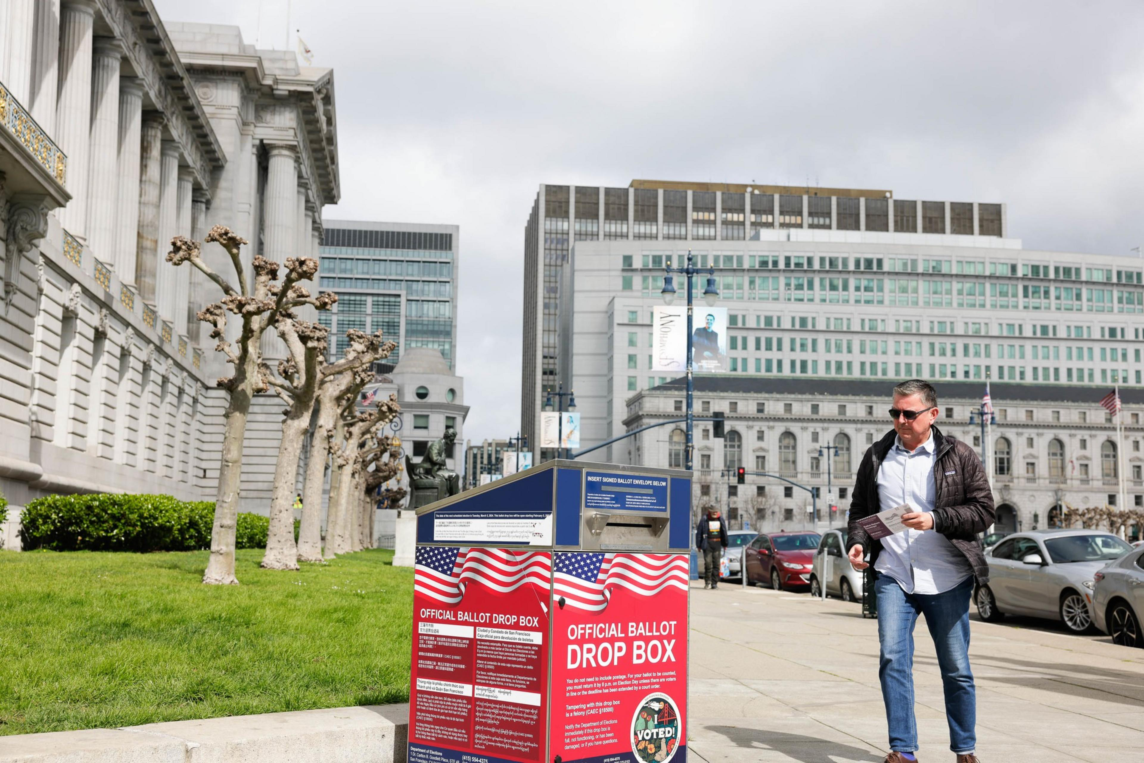 A person walks past a ballot drop box outside a building with classical architecture under a cloudy sky.
