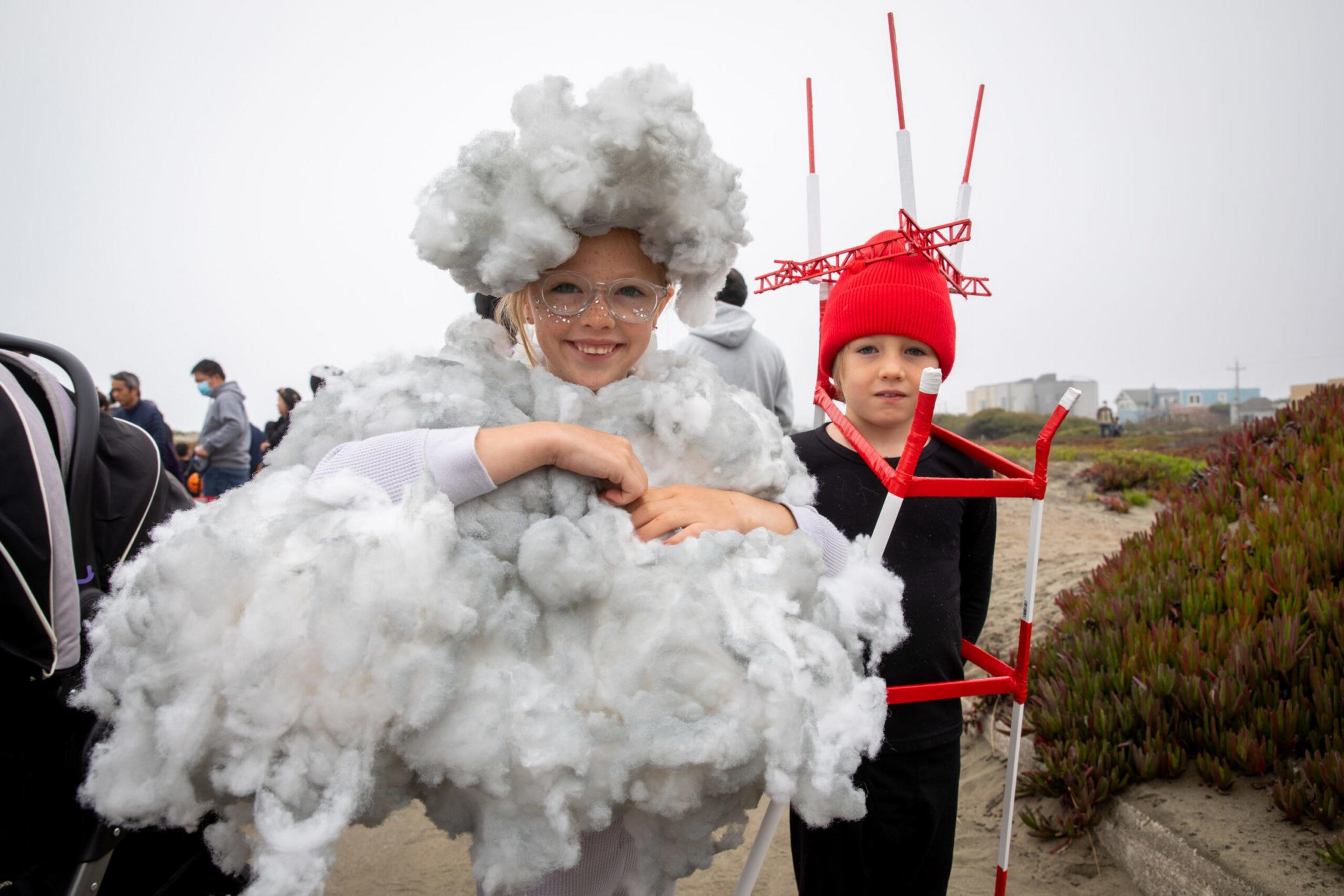 A child dressed as a fluffy gray cloud stands next to another child wearing a red hat with red and white structures resembling radio towers.