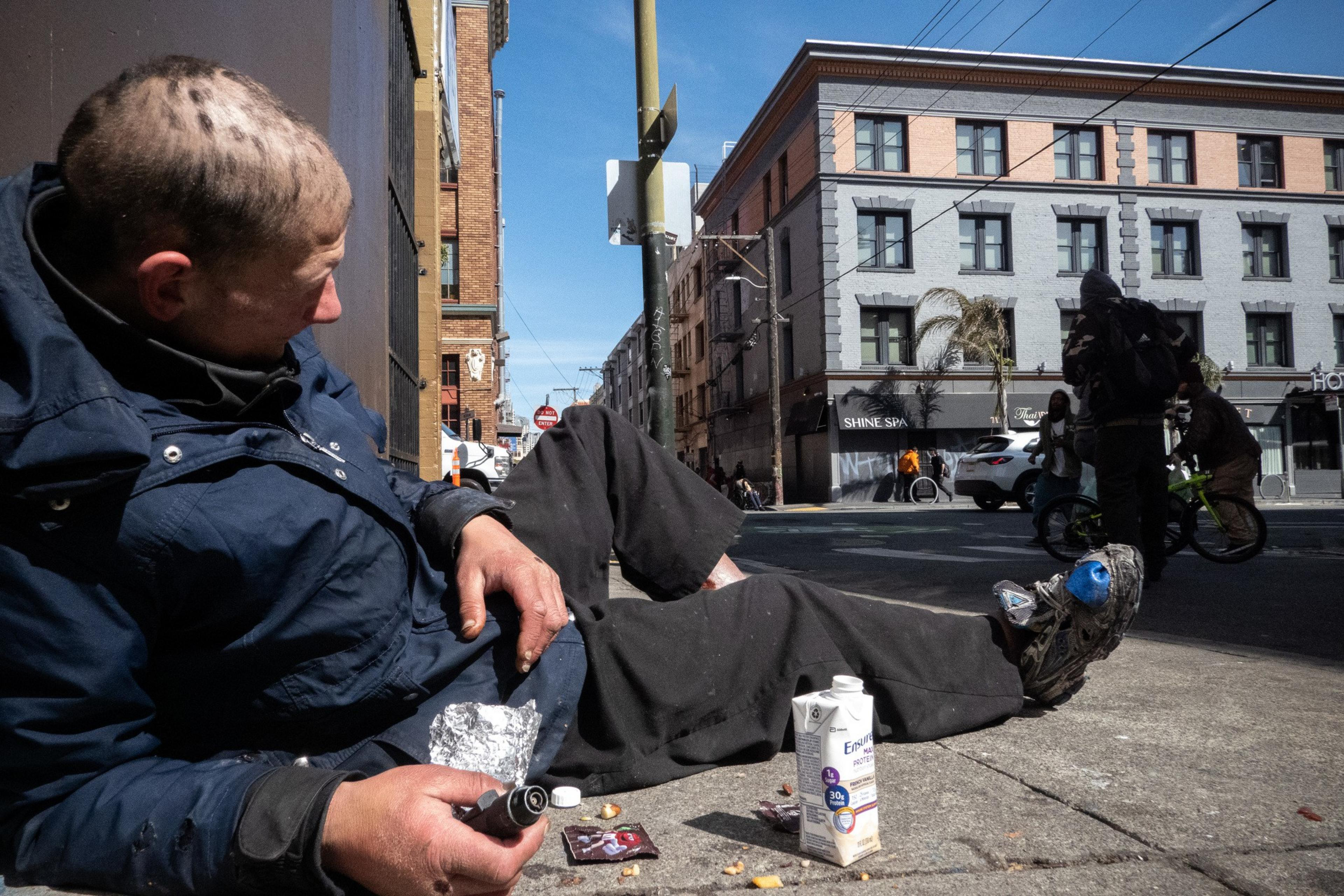 A person sitting on a sidewalk with items scattered around, amidst an urban backdrop.