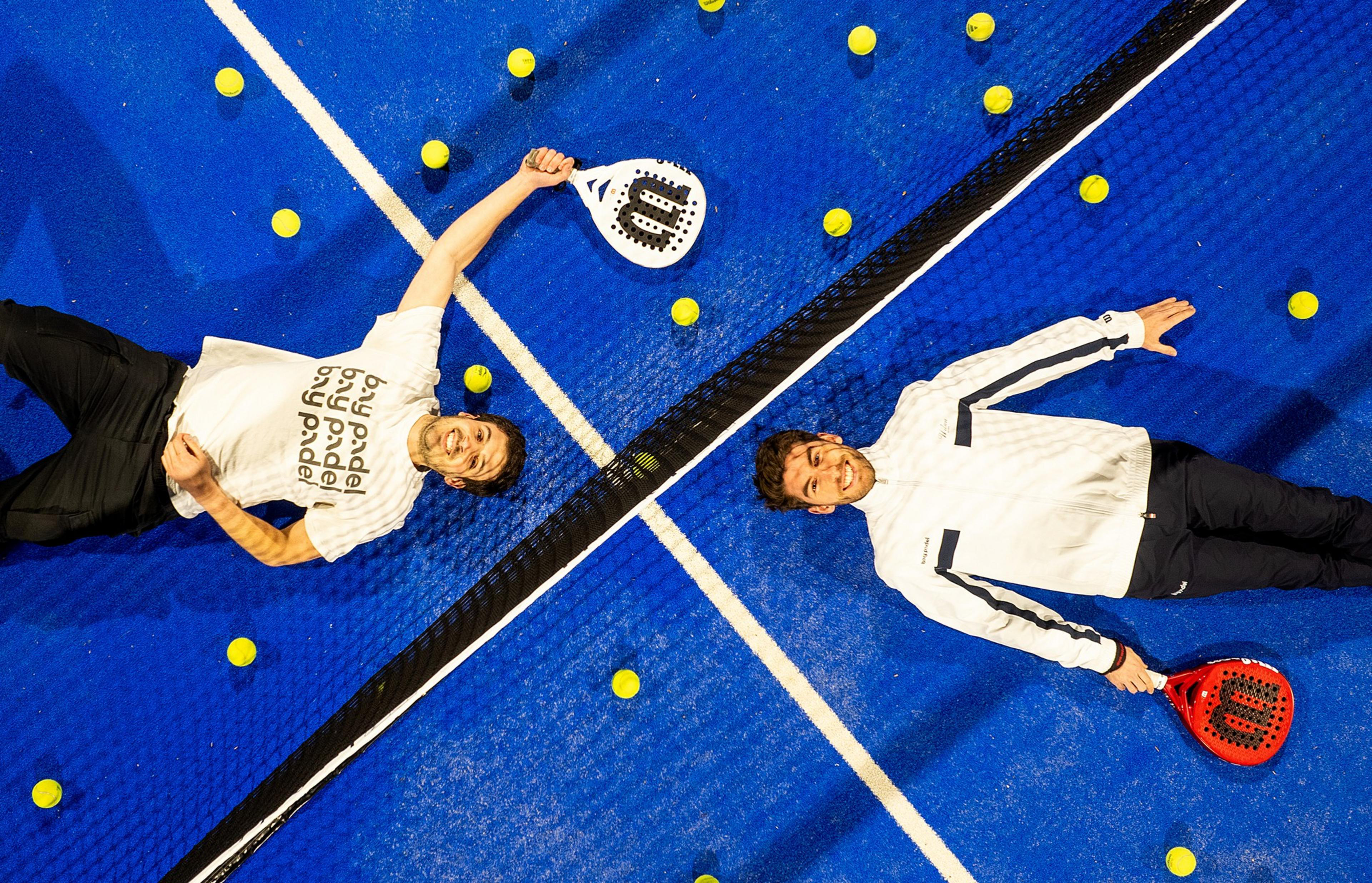 Two men in sportswear lie on a blue padel court, smiling and holding rackets. Yellow balls are scattered around, and a net divides the court.