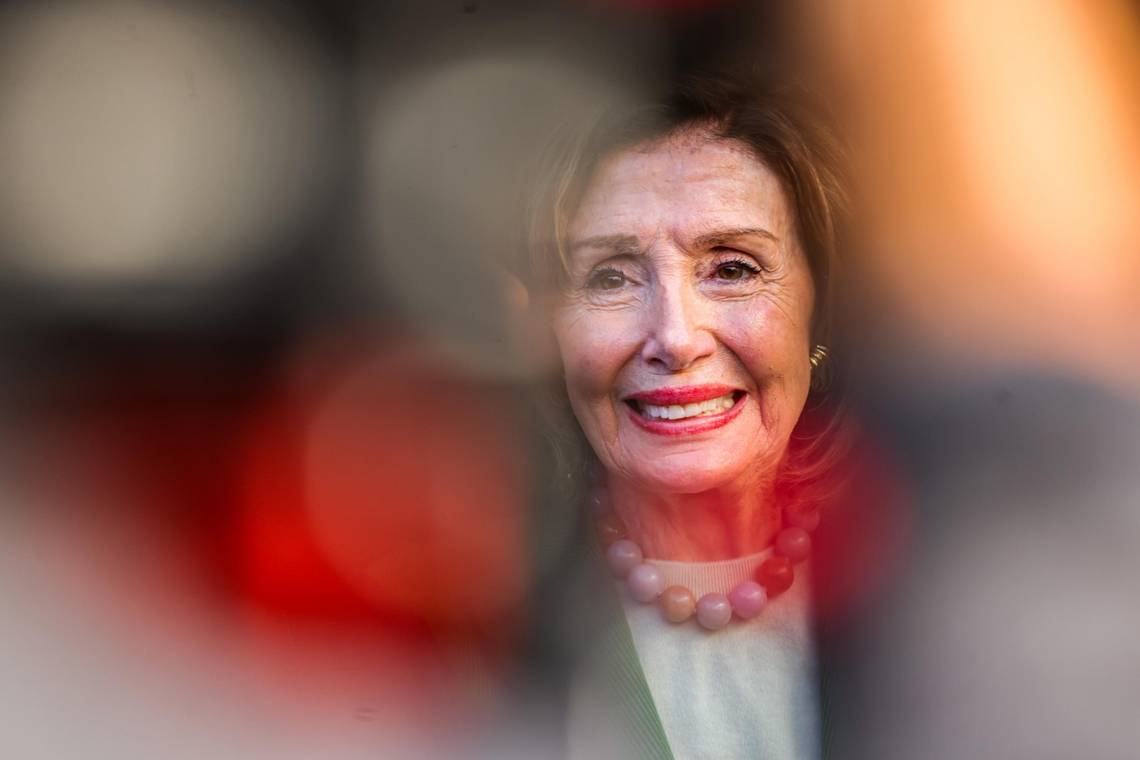 A smiling woman with light skin, wearing a pink beaded necklace and earrings, is seen through blurred, out-of-focus objects creating a soft, colorful frame.