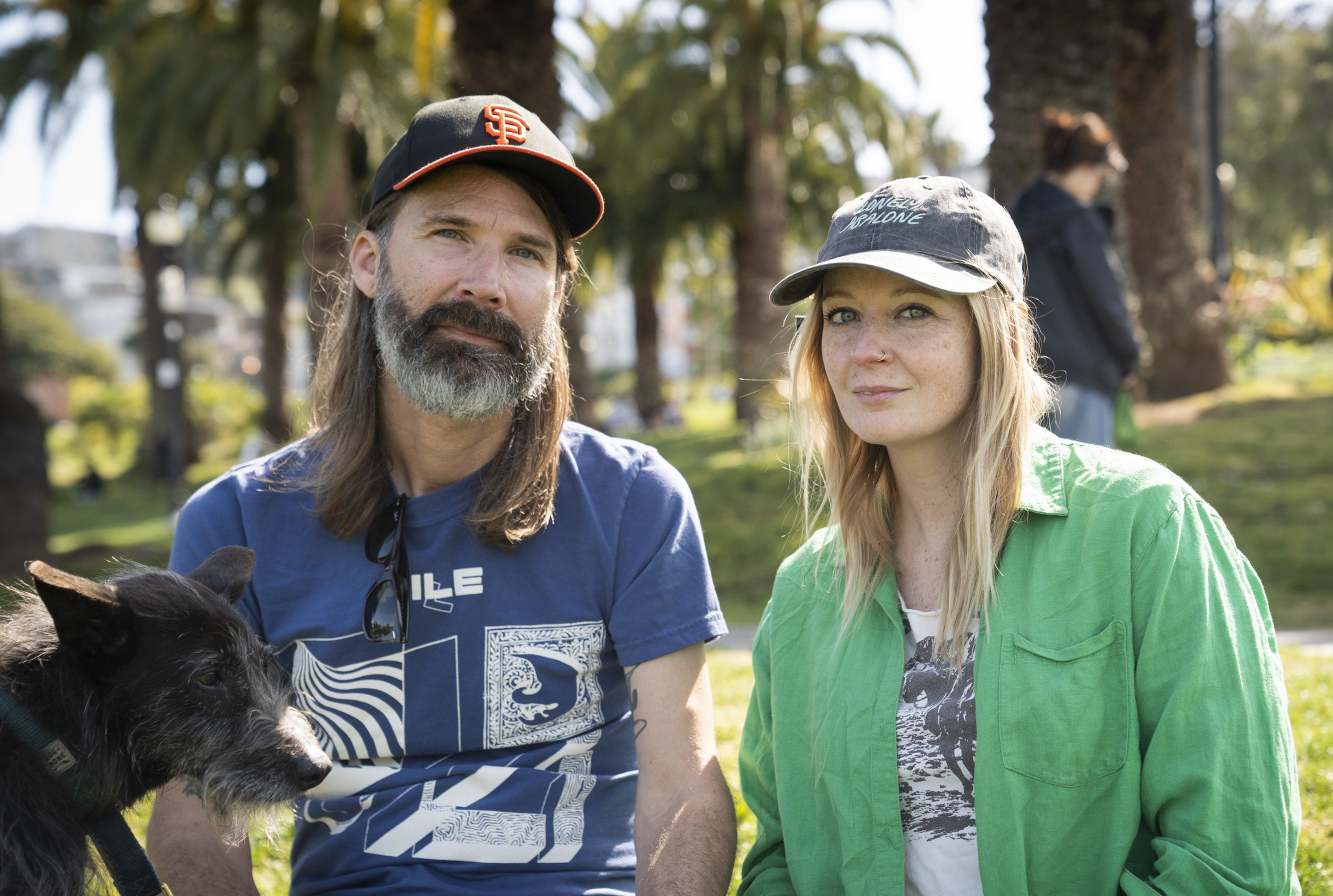 A man with a beard and a cap and a woman with freckles in a green shirt, both sitting outside with a black dog nearby. Palm trees are in the background.