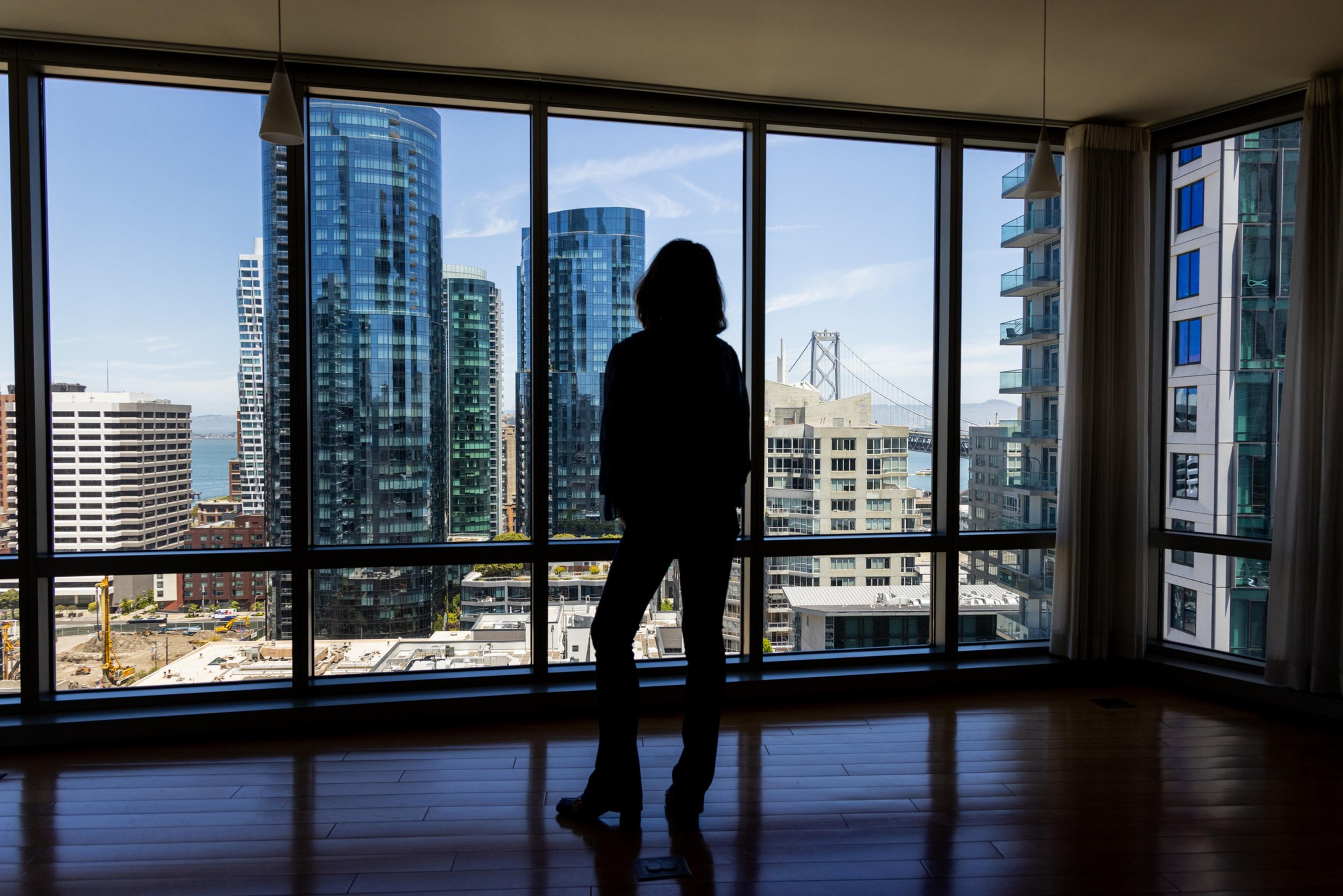 A person stands inside, silhouetted against floor-to-ceiling windows, overlooking a cityscape with tall, modern buildings and a bridge in the background.