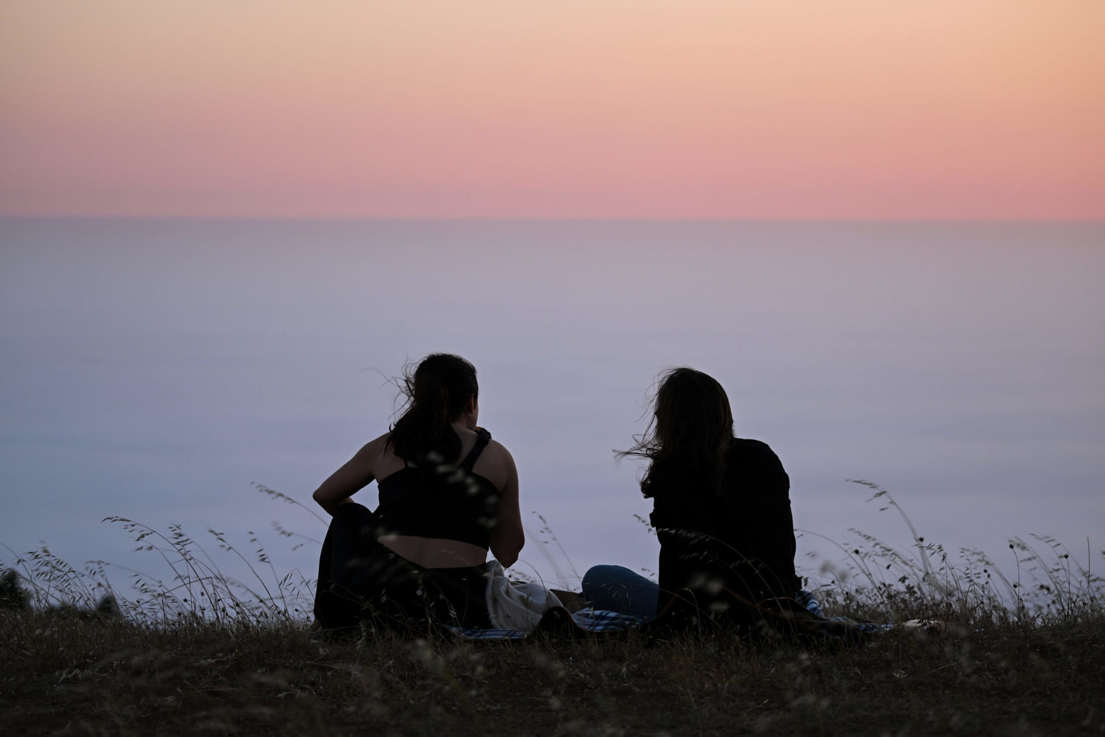Two people sit on a grassy hill facing a calm sea under a soft pink and purple sunset sky.