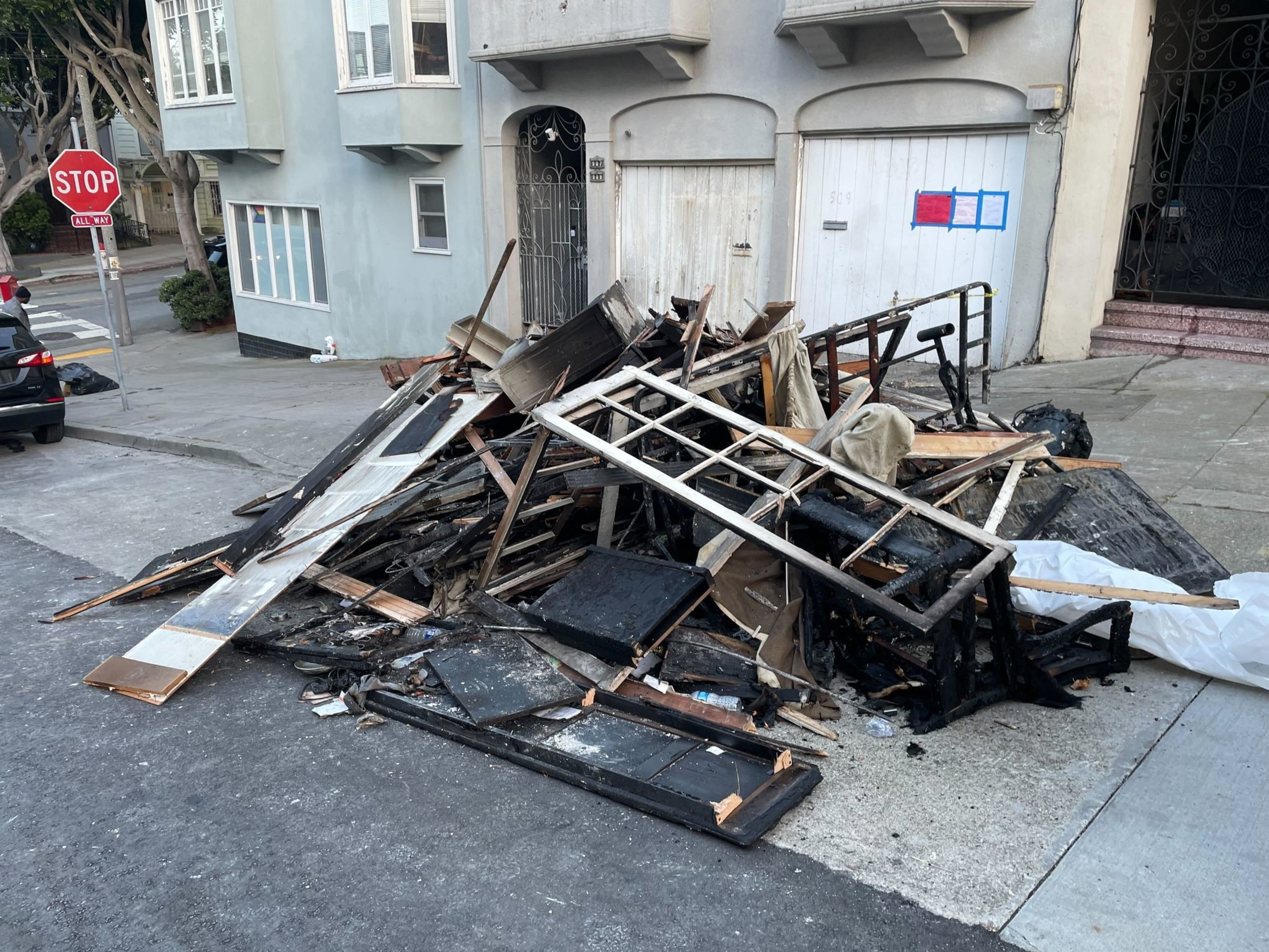 A heap of charred wooden debris and furniture lies on a city street corner near pale green and white buildings with a stop sign visible.