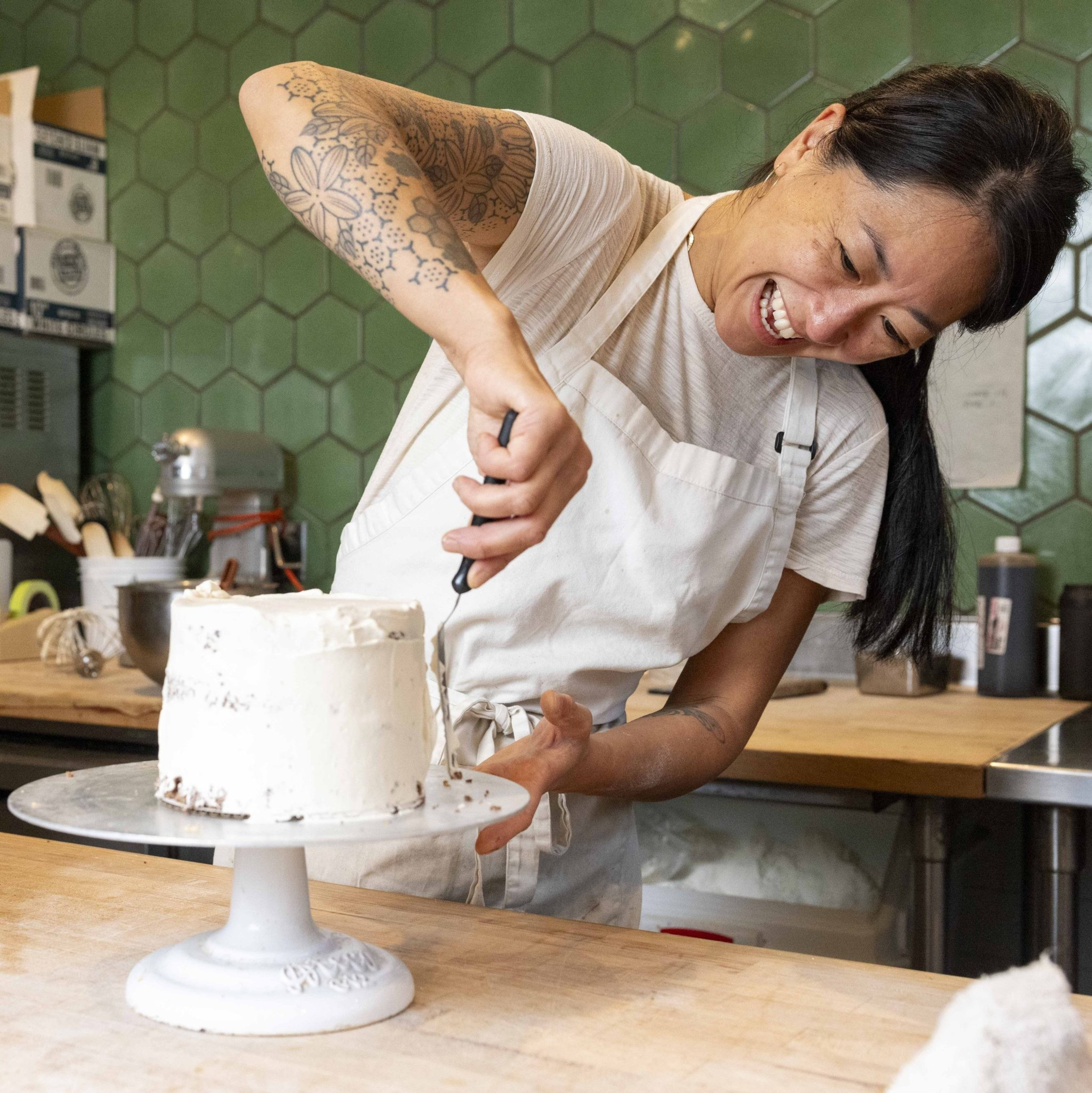 A joyful person with long hair decorates a white frosted cake on a stand. They're in a kitchen with green hexagonal tiles and kitchen supplies around.