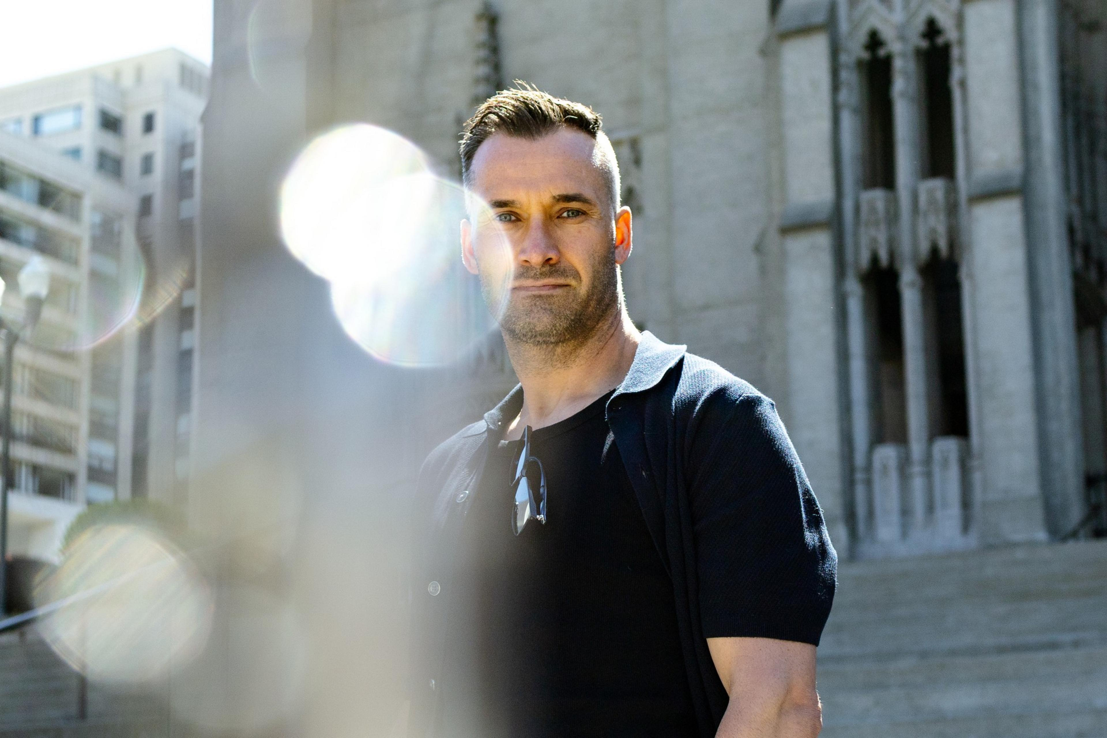 A person stands in front of a grand building, possibly a cathedral. They're wearing a dark shirt, and the image has bright, blurred light spots.