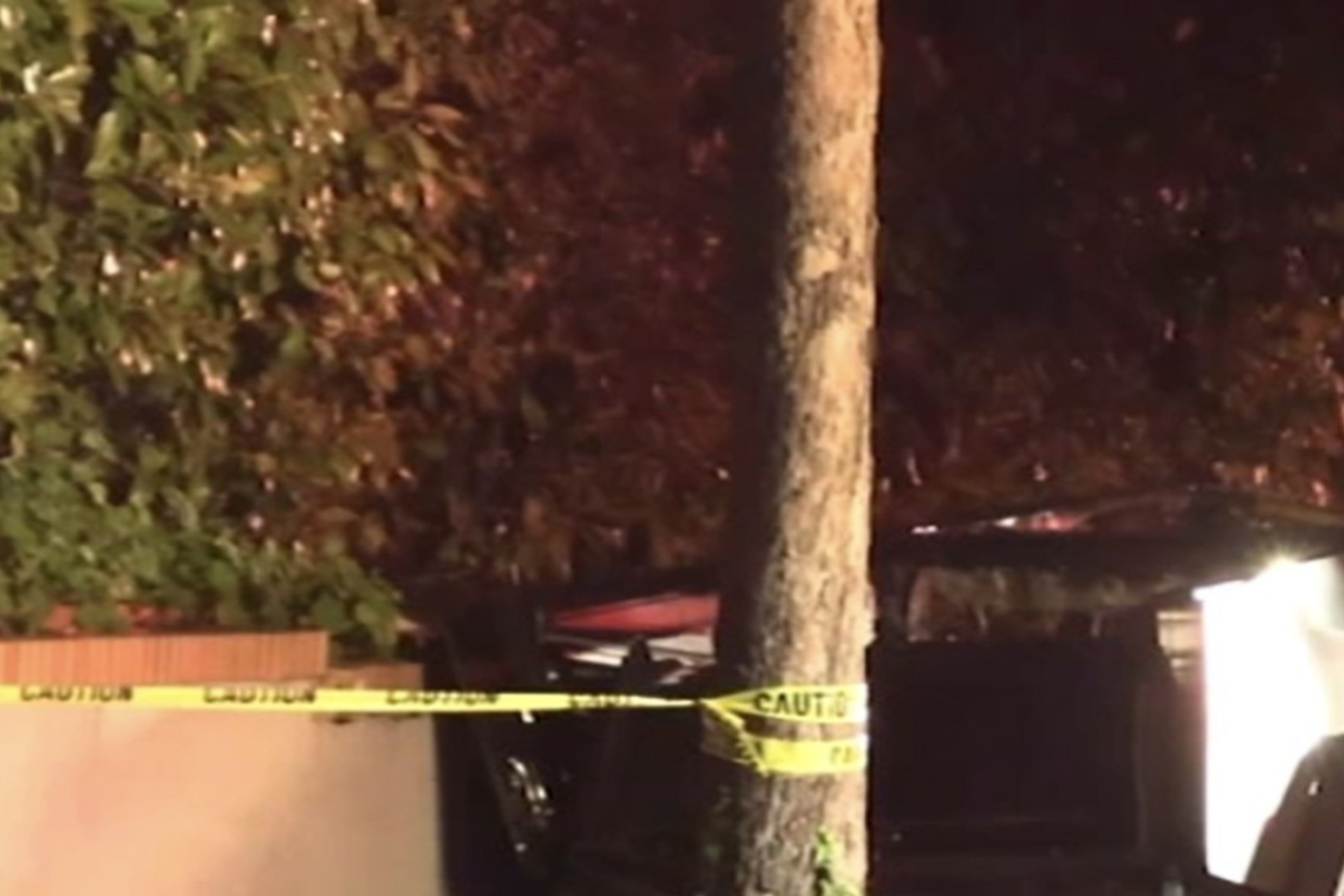 A damaged car is crashed into a tree beside a wall, surrounded by caution tape. It appears to be nighttime, with some debris on the ground.