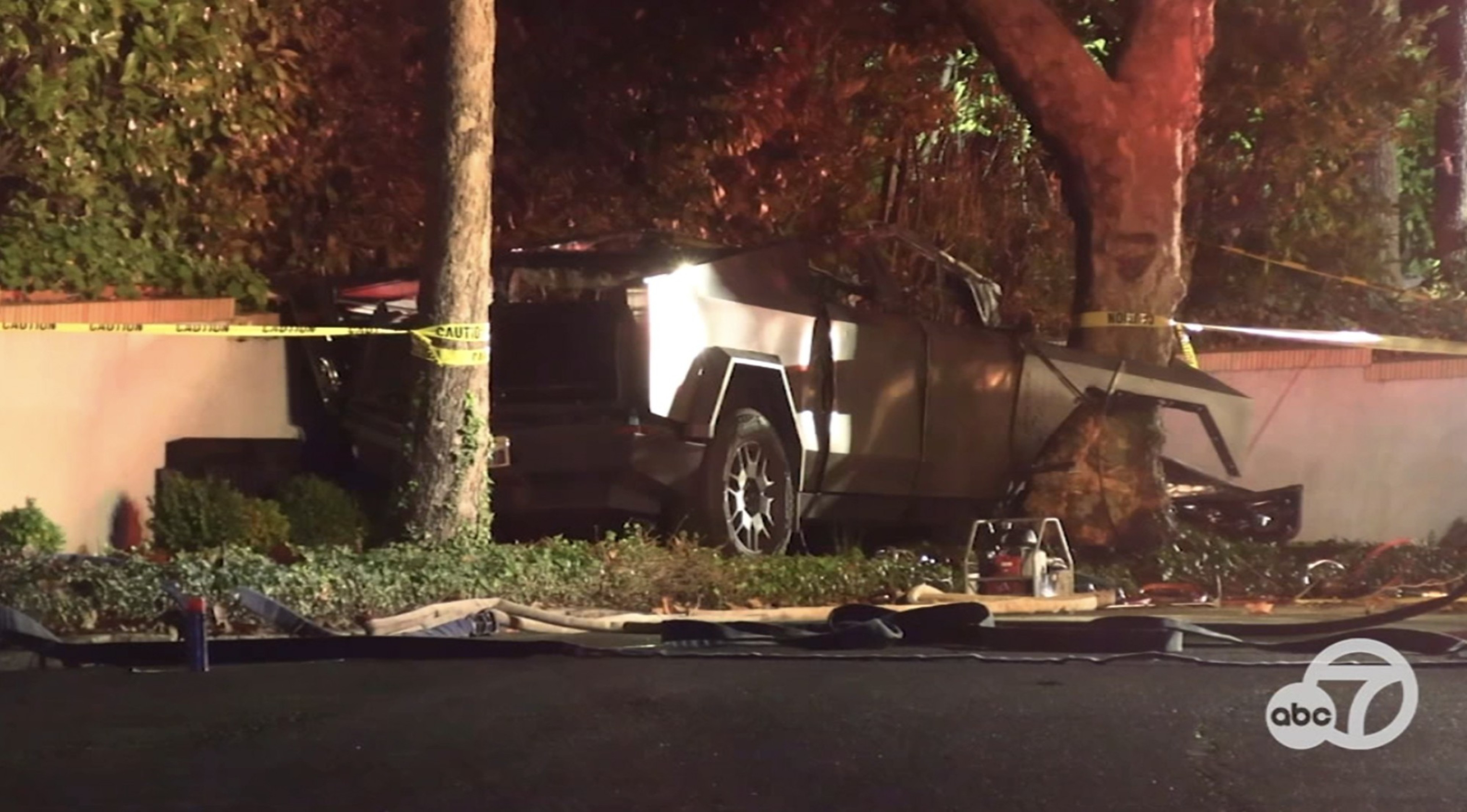 A damaged car is crashed into a tree beside a wall, surrounded by caution tape. It appears to be nighttime, with some debris on the ground.