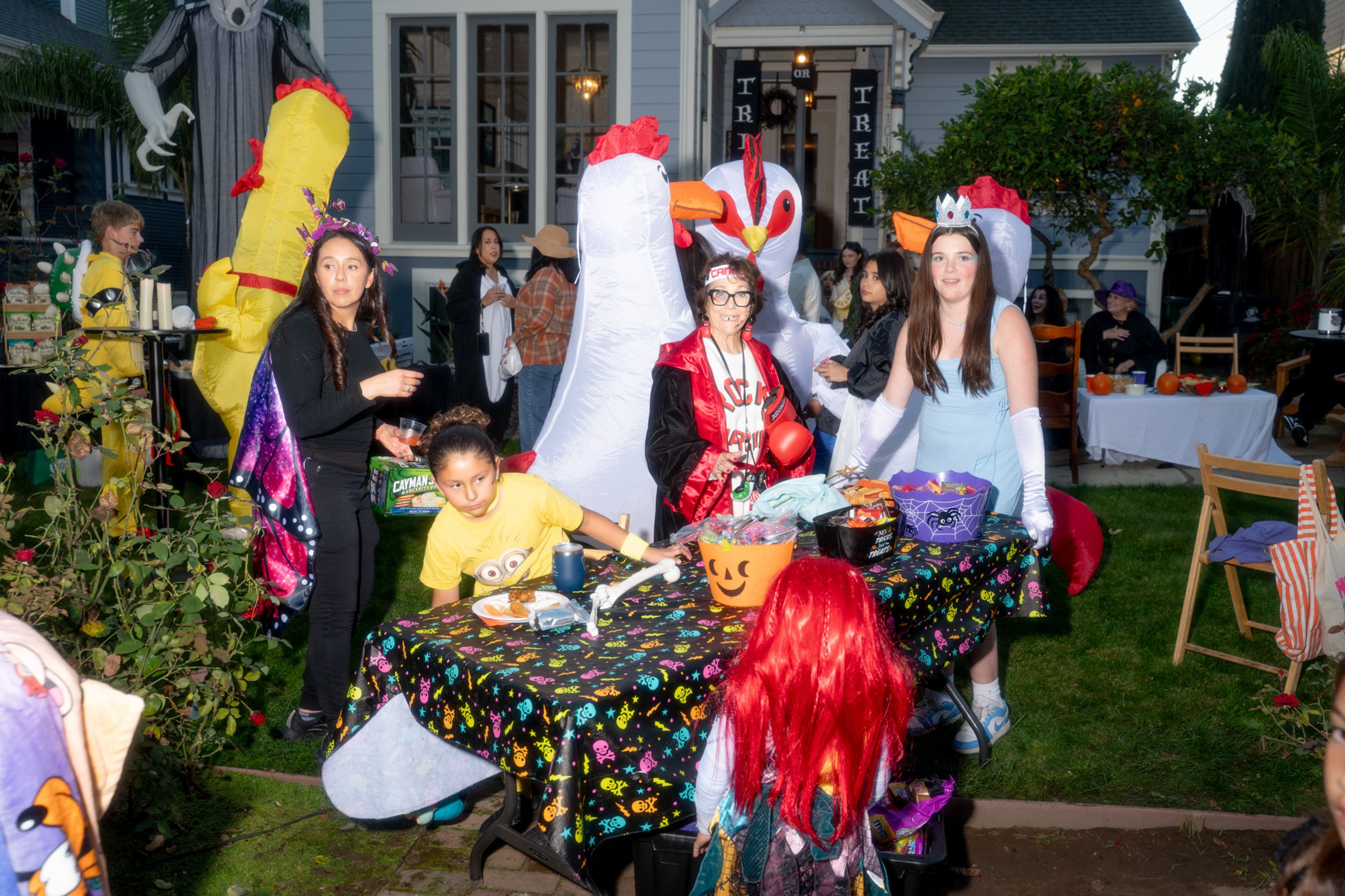 People in colorful costumes, including inflatable chickens, gather around a decorated table with Halloween candy buckets in a lit backyard at dusk.