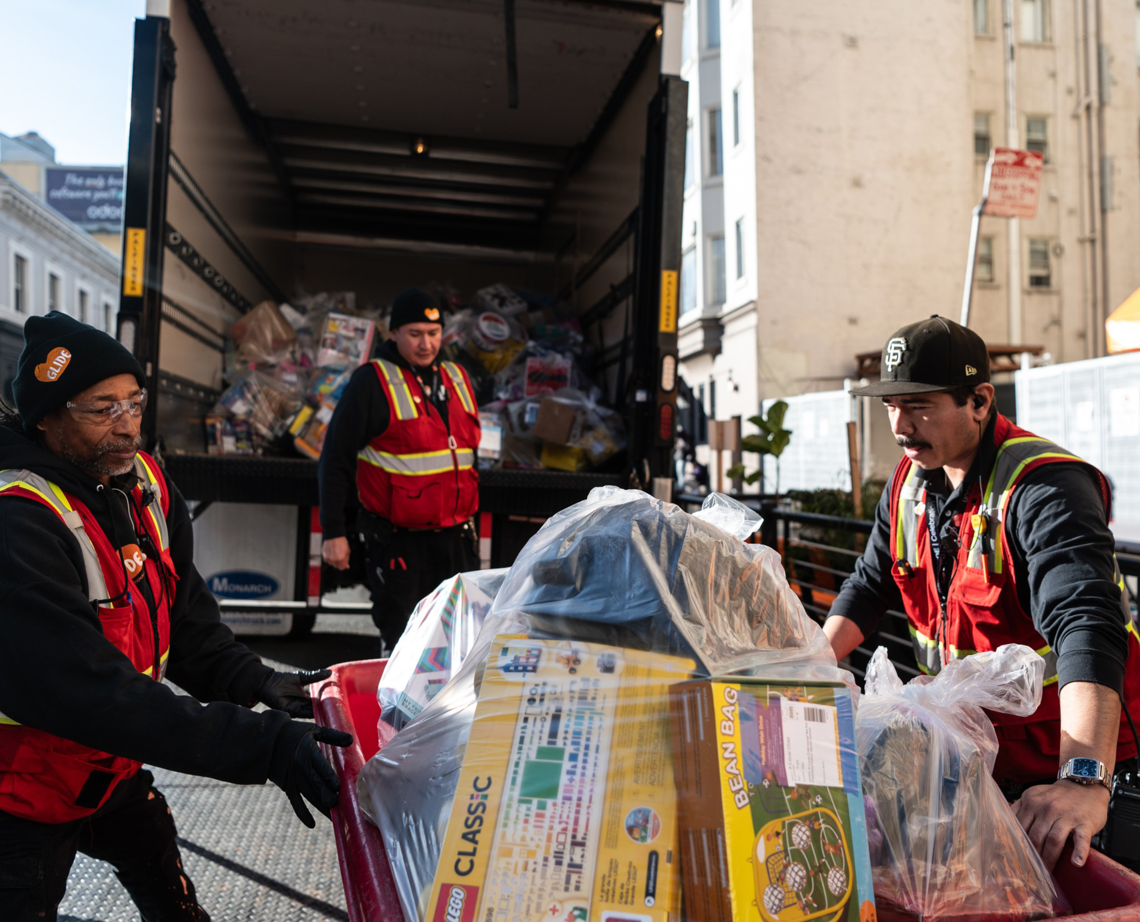 Three people in red safety vests unload toys from a truck. One person is pushing a cart with boxes, including a LEGO set and a bean bag.