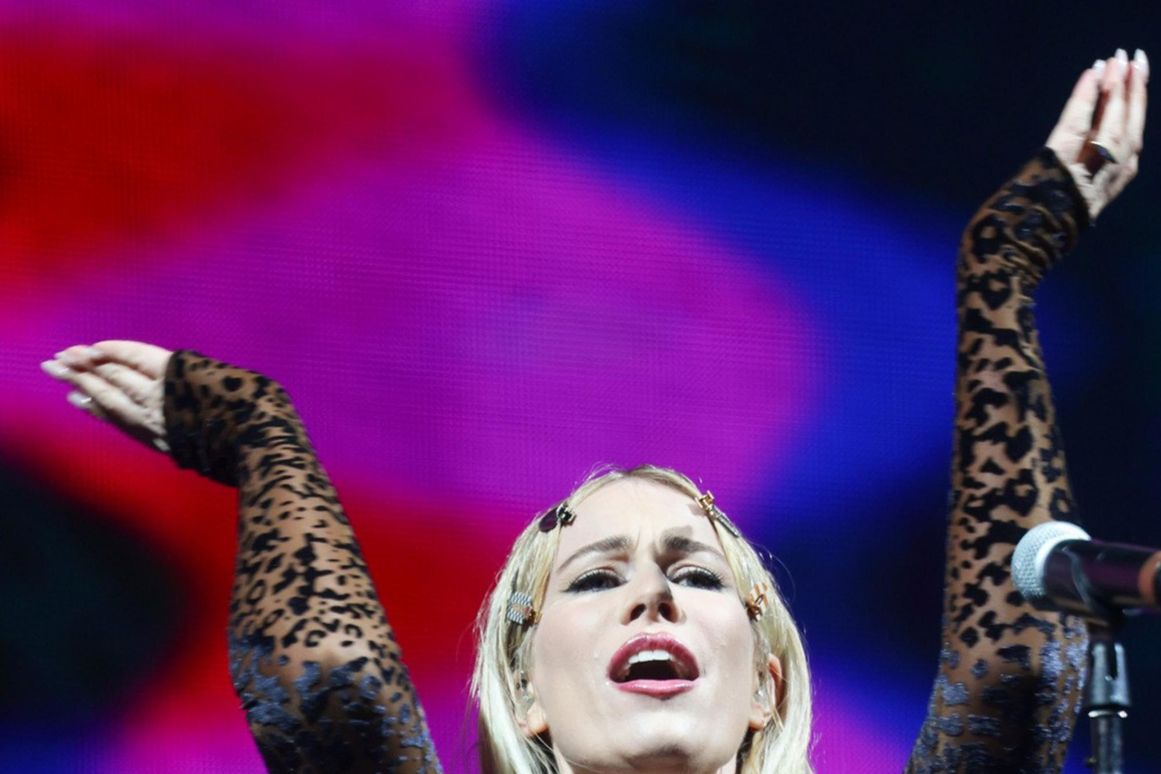 A person wearing a leopard-print jumpsuit performs on stage, with arms raised and a microphone nearby. The background is a colorful, blurred light display.