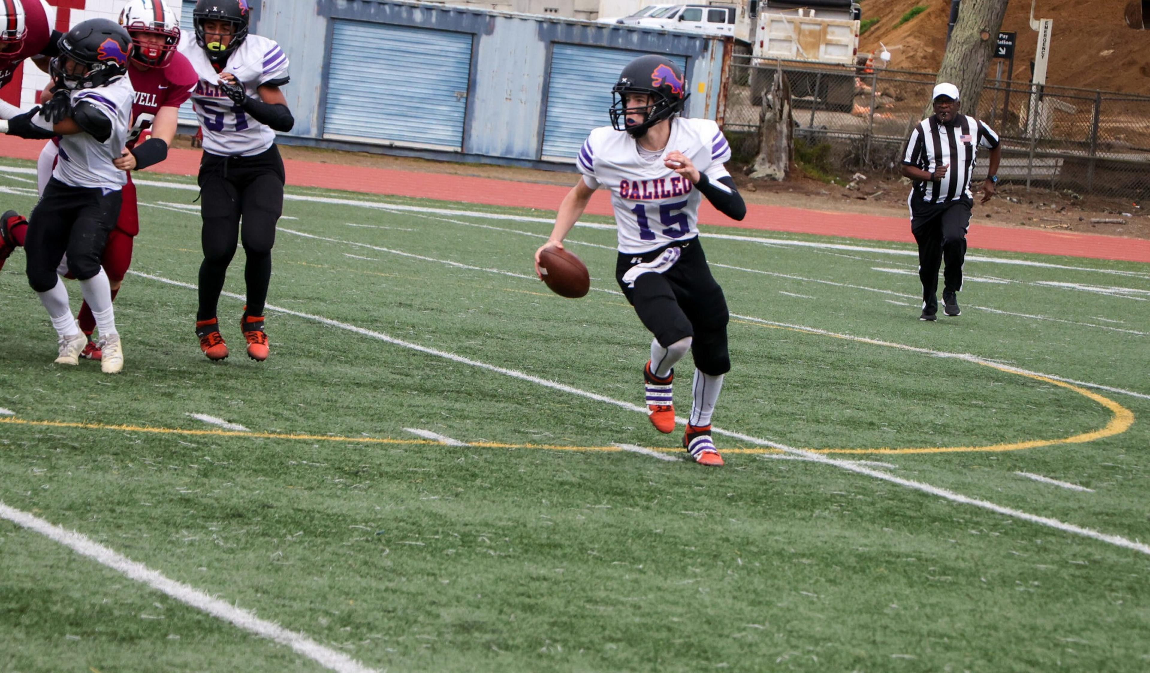 A football player in a white and purple uniform runs with the ball, while opponents and a referee run behind him on the field.