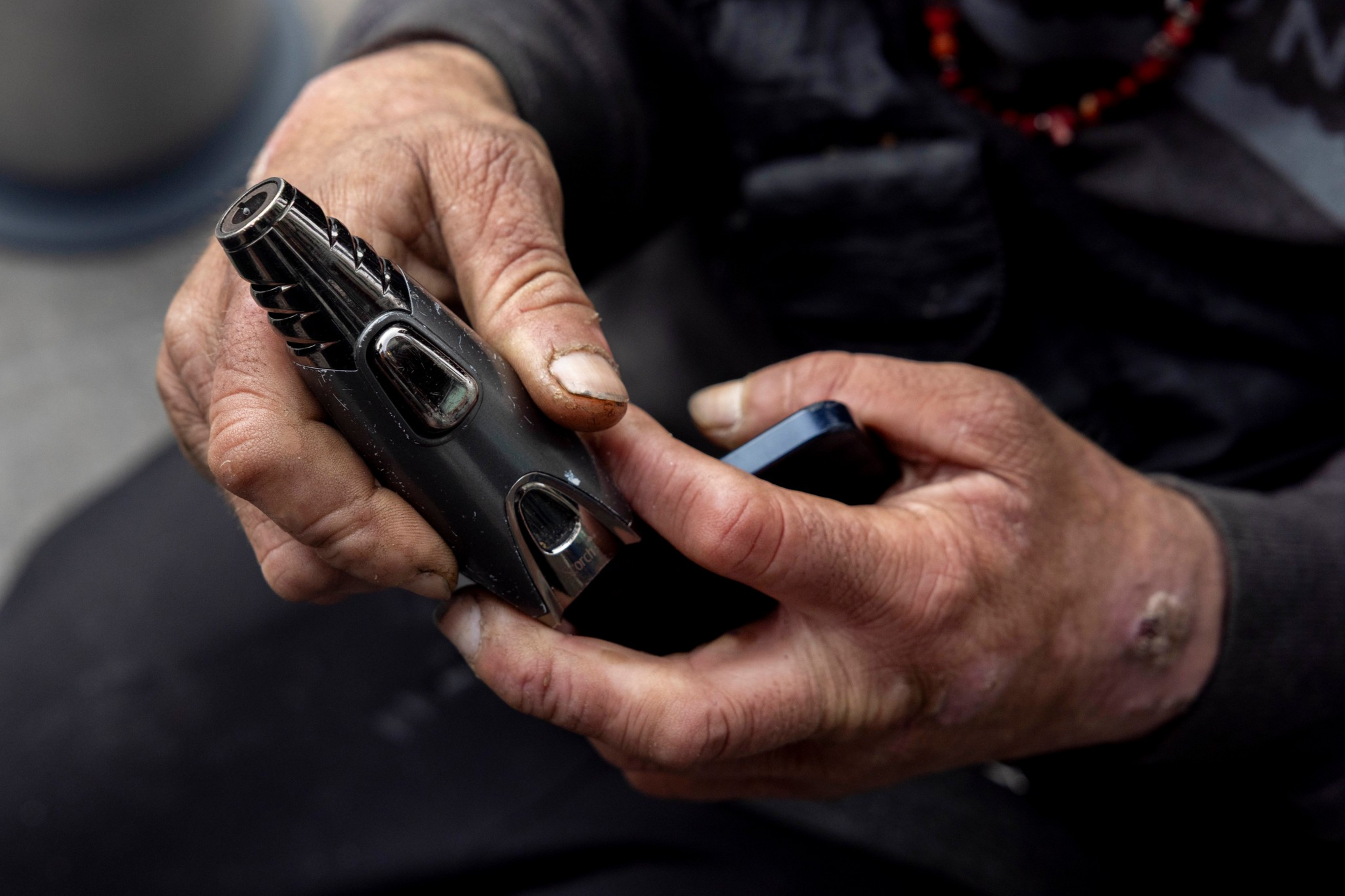 A person with rough, worn hands holds and operates a small black electronic device, possibly a vape or a lighter.