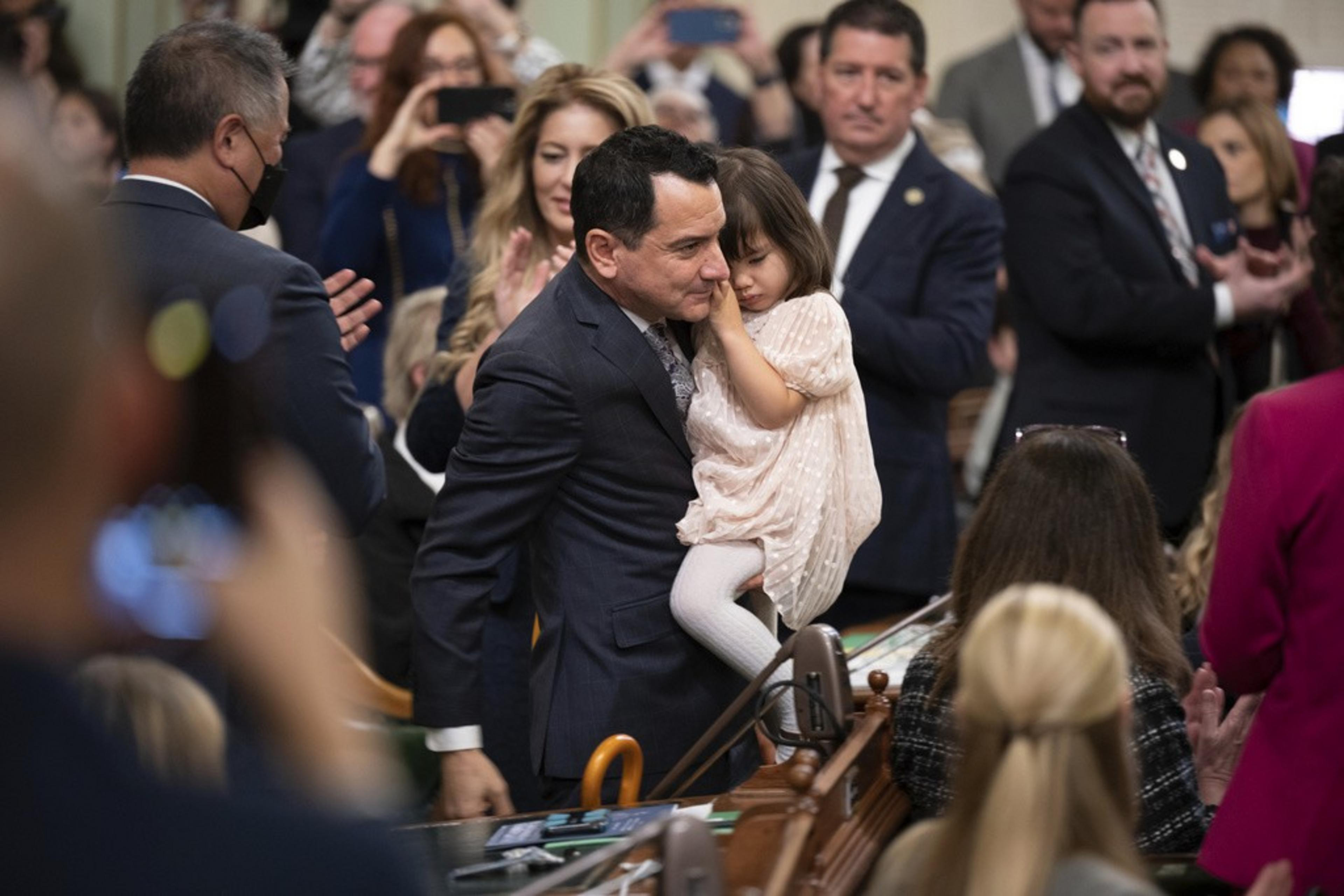 A man in a suit holds a young girl amidst a crowd of people, many clapping or taking photos.
