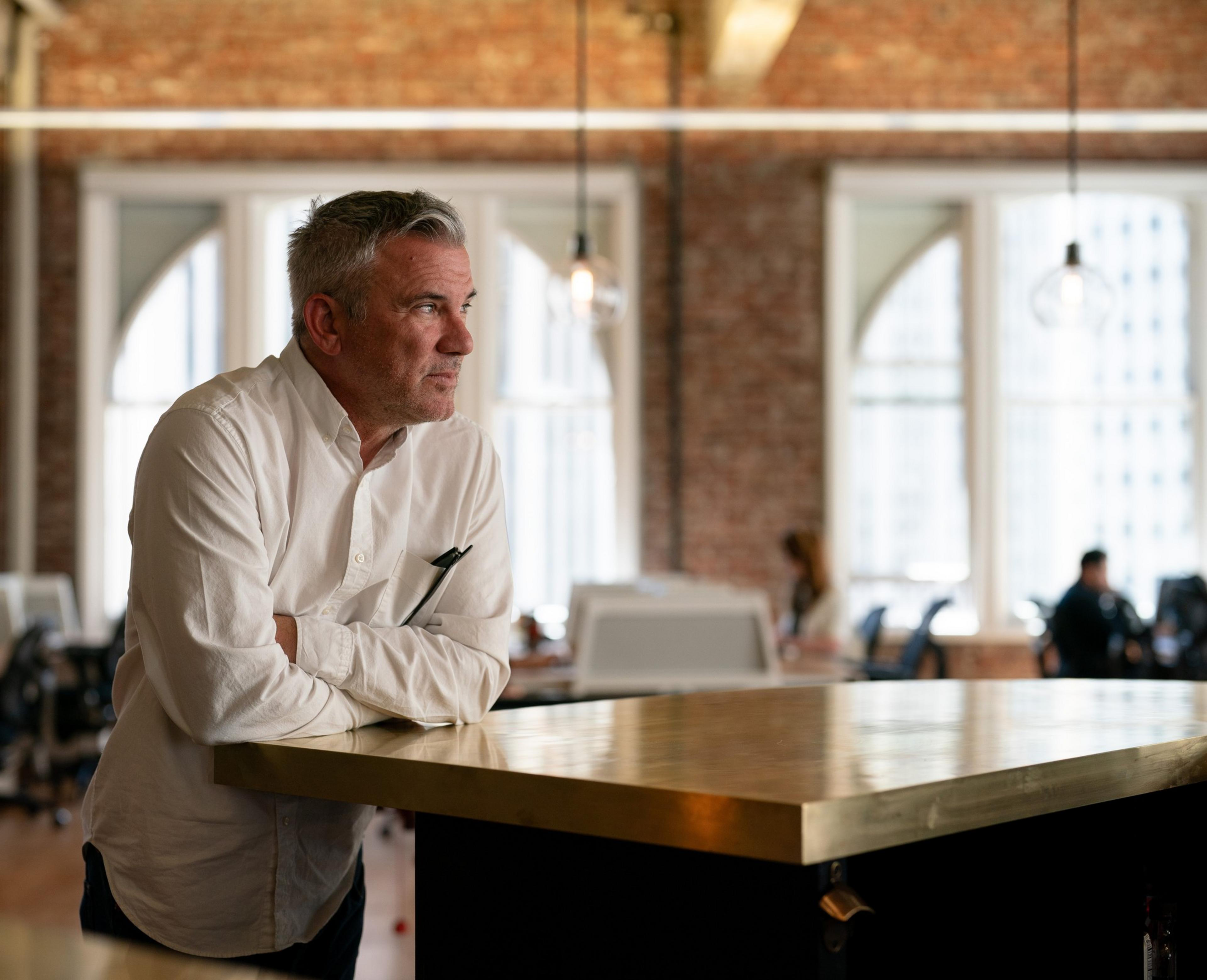 A man in a white shirt leans on a counter in an industrial-style office with brick walls and arched windows. Several people work in the background.