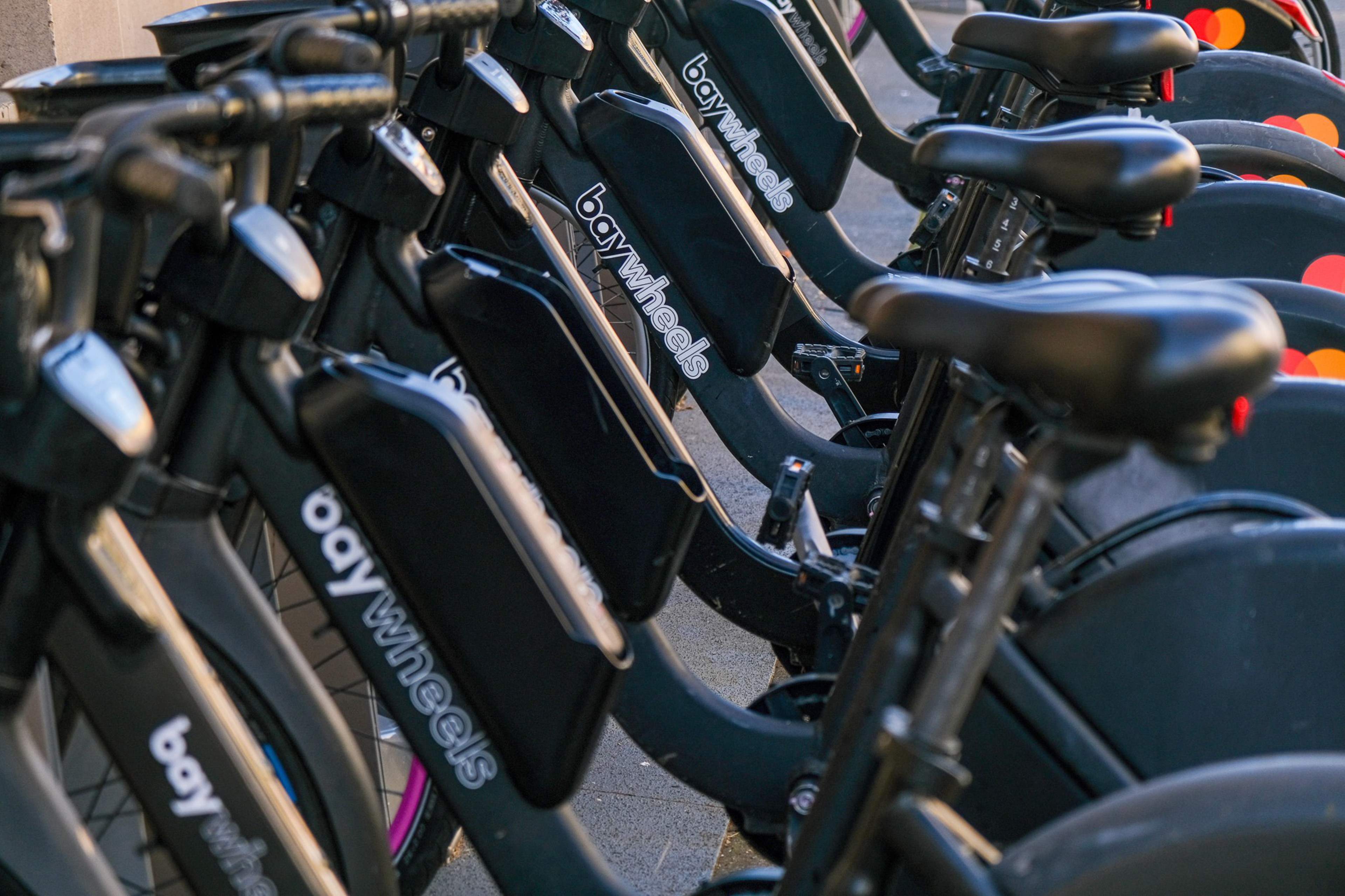 A row of black electric bikes with "bay wheels" branding is lined up, showing their seats, handlebars, and battery compartments.