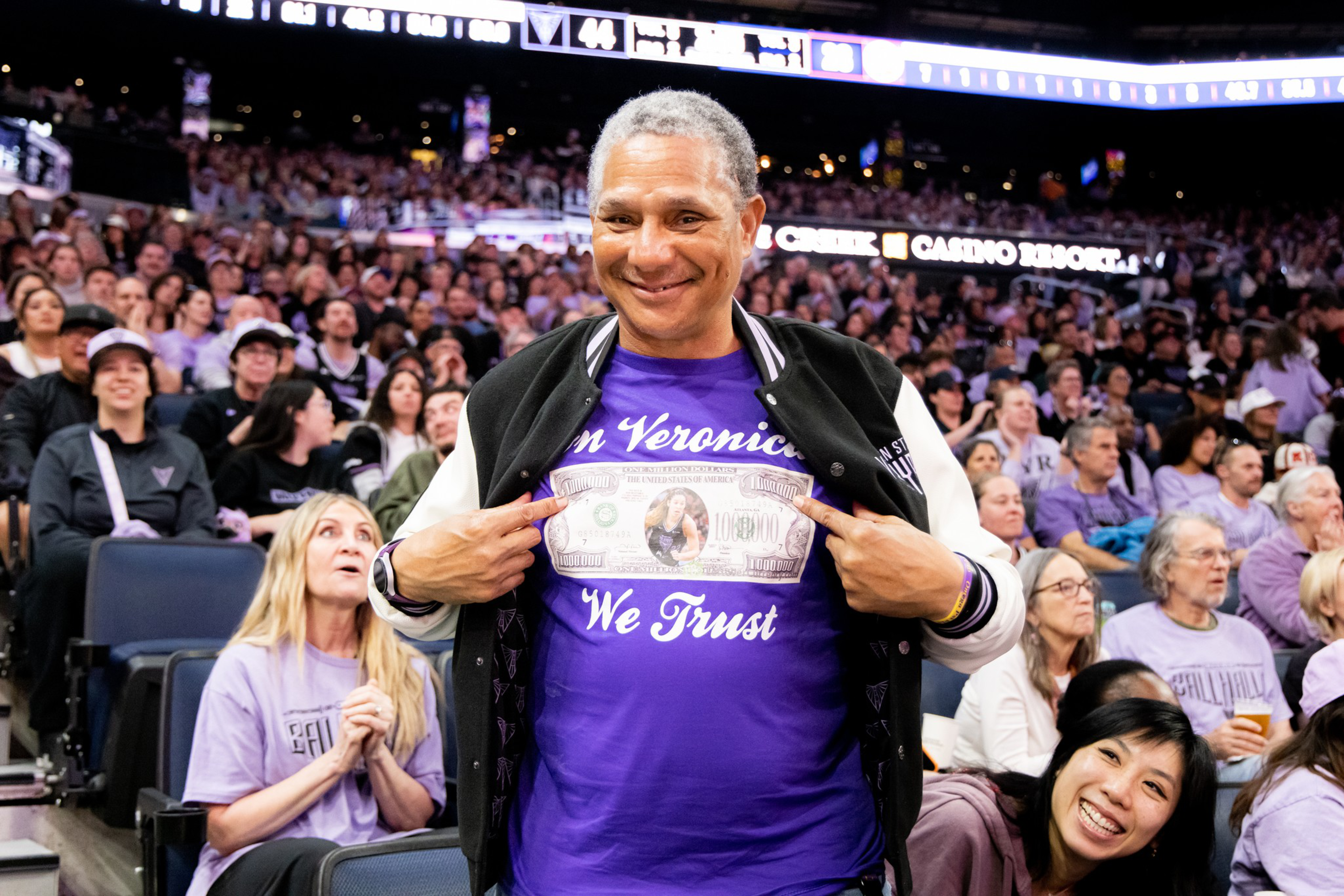 A man smiles and points at his purple shirt that reads “In Veronica We Trust” with a dollar bill design featuring a woman’s image, surrounded by a crowded arena.