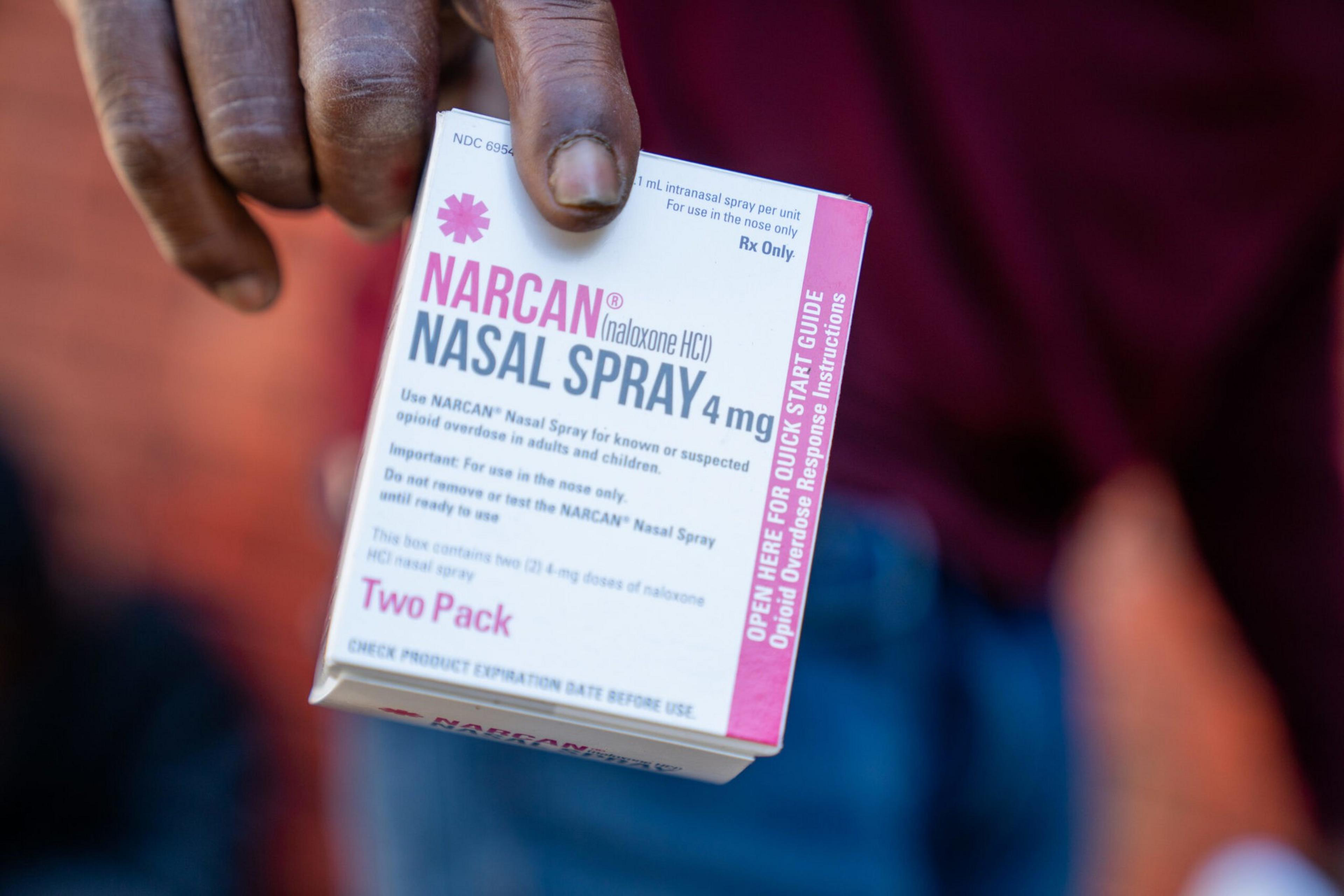 A hand holds a box of Narcan Nasal Spray 4 mg, used for opioid overdose, labeled for intranasal use with a "Two Pack" and quick start guide.