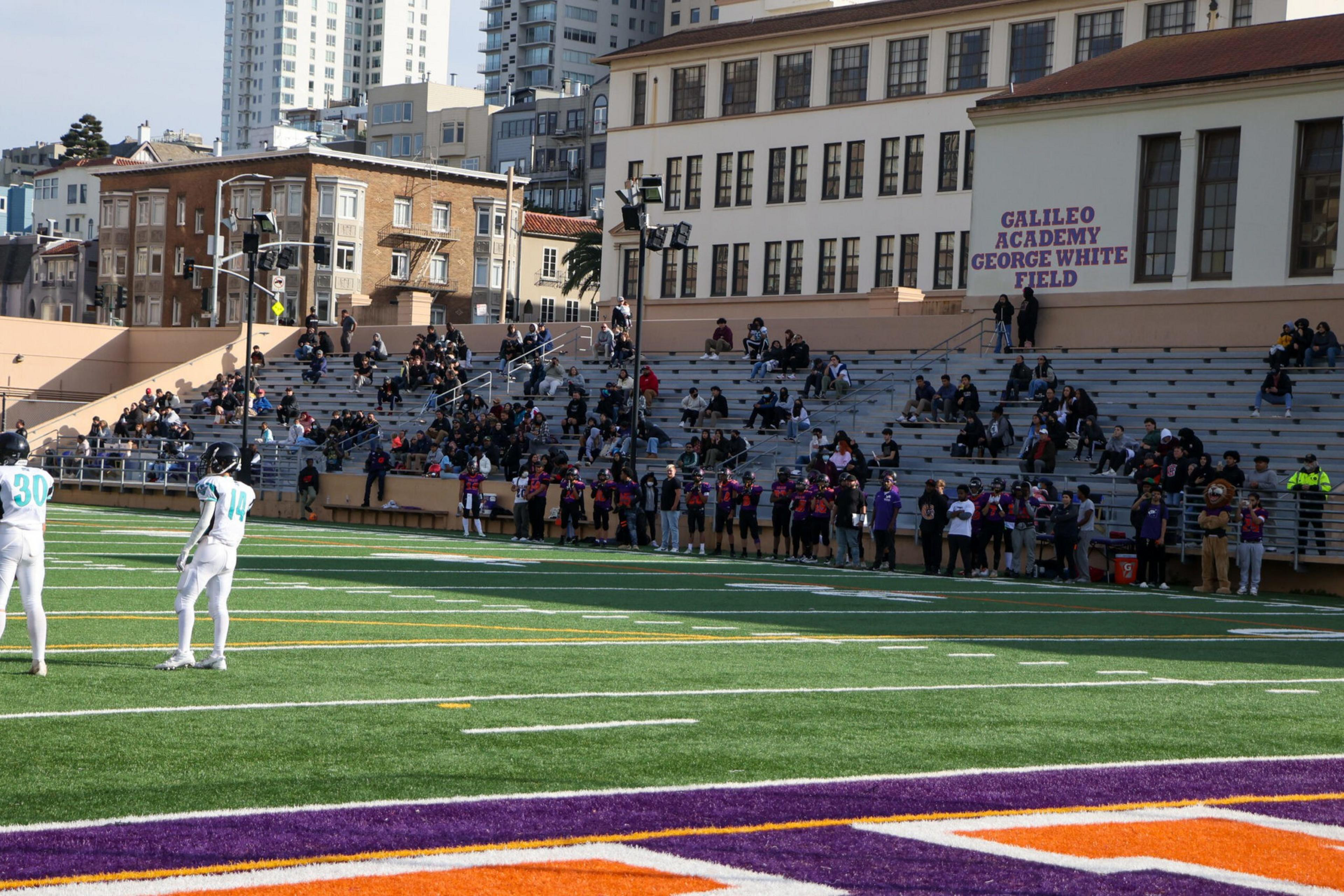 A football field with two players in white uniforms stands in the foreground, while spectators sit in bleachers near a building labeled "Galileo Academy George White Field."