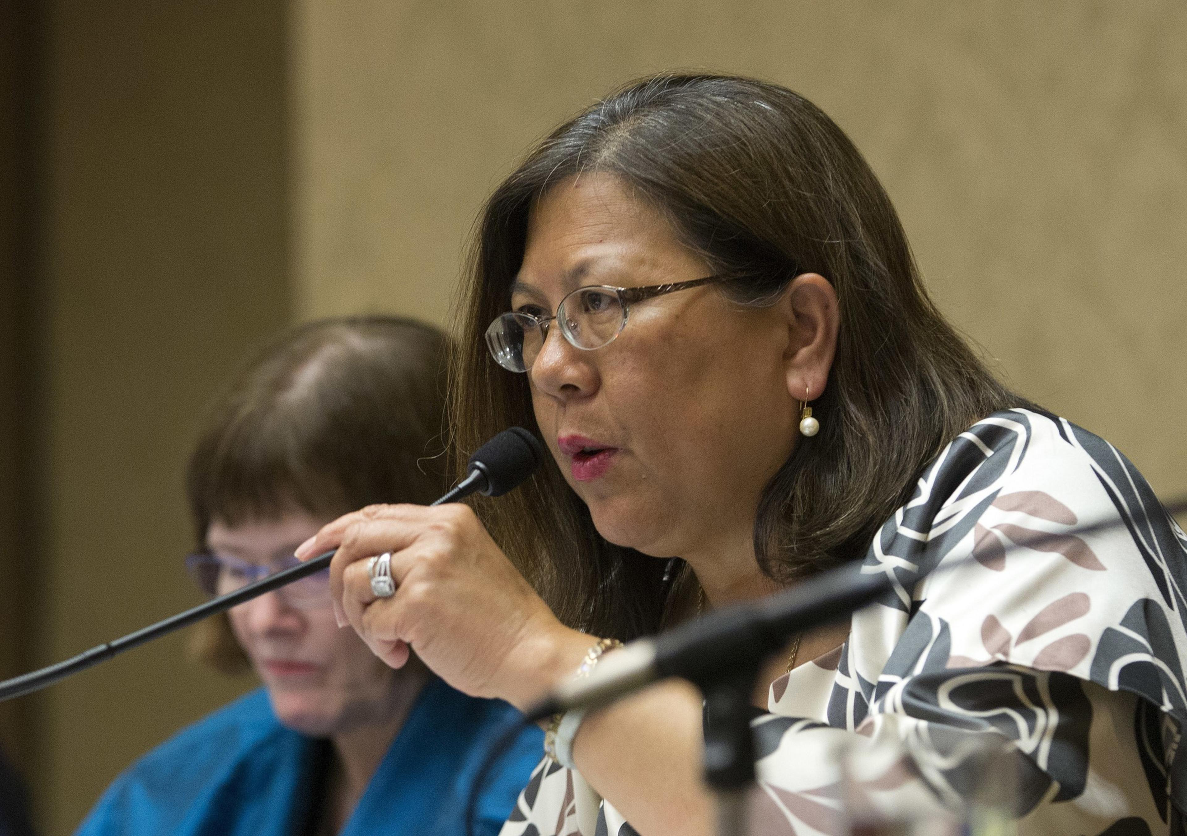 A woman speaks into a microphone, wearing glasses and a patterned blouse, with another person slightly visible behind her.