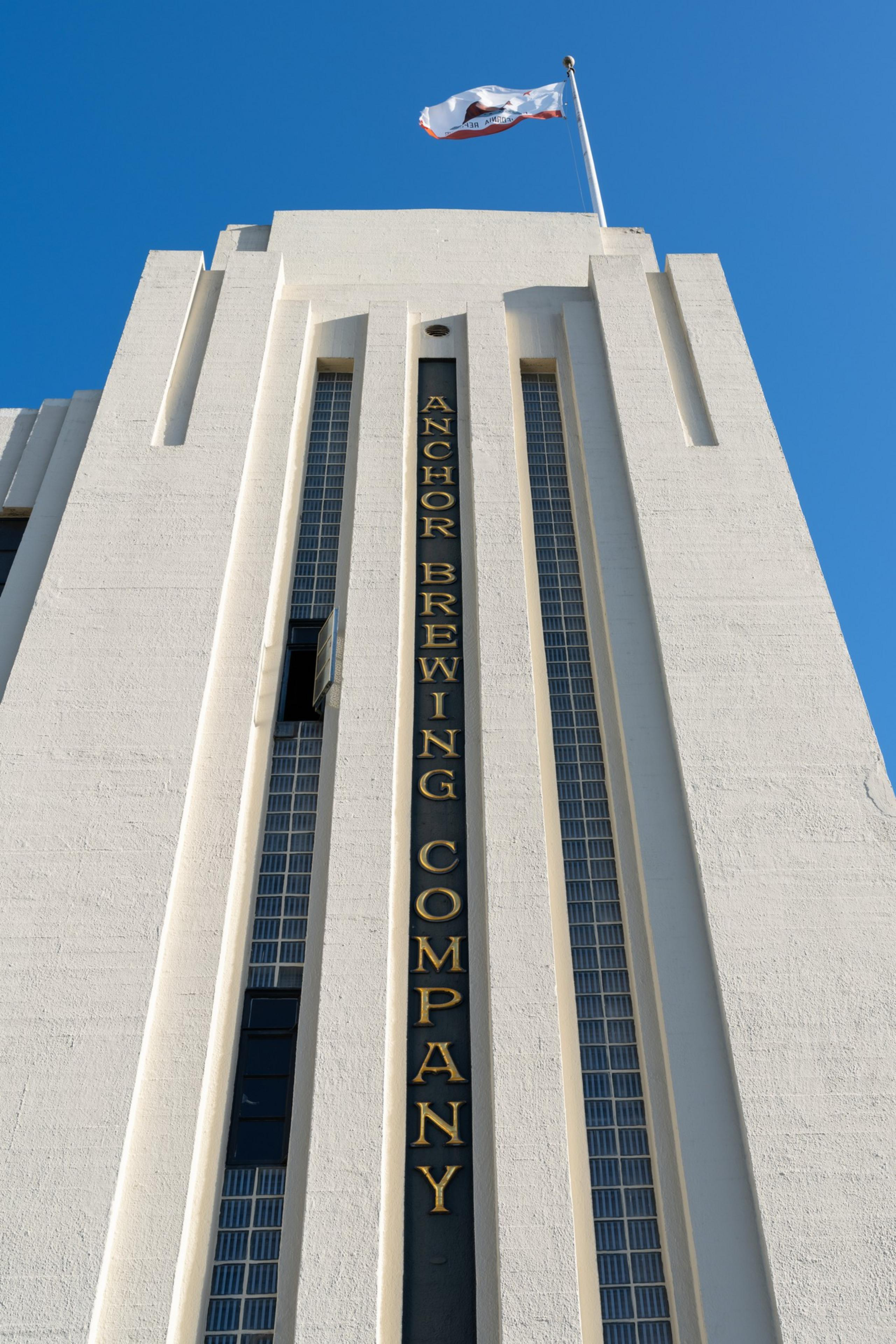 A tall white building with vertical windows displays &quot;Anchor Brewing Company&quot; in gold letters, topped with a waving flag against a clear blue sky.