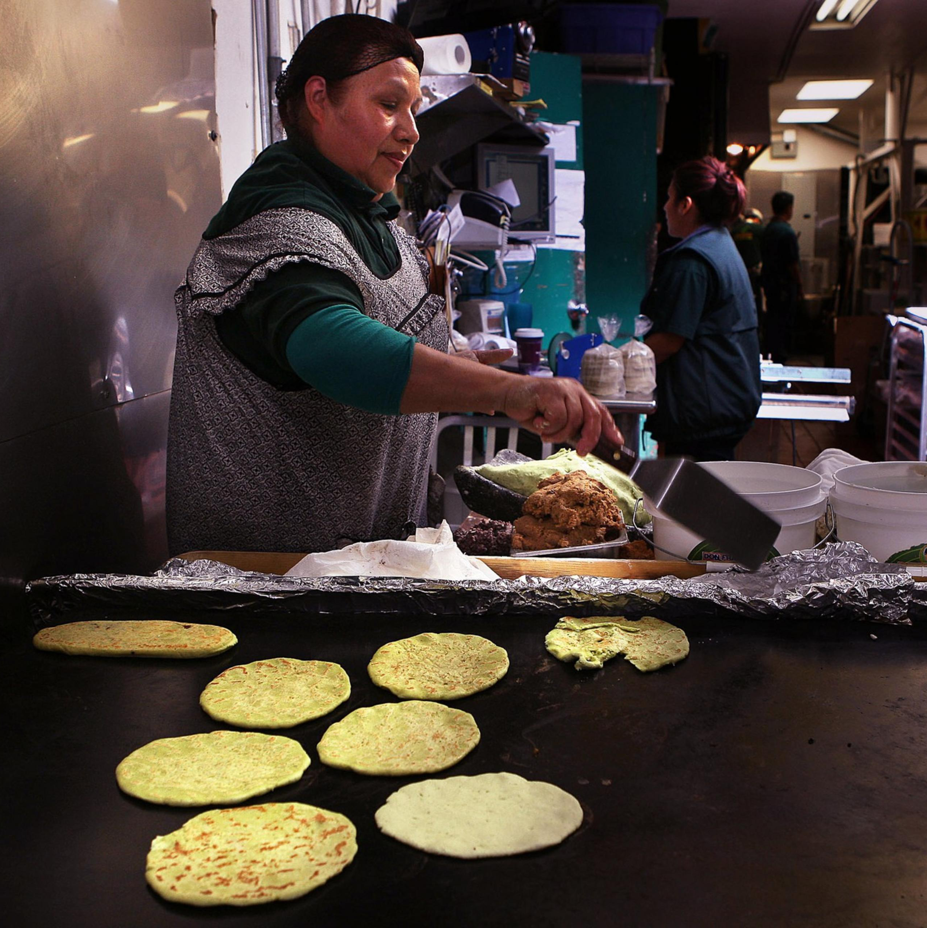 A woman in an apron cooks tortillas on a large griddle in a kitchen. Several people are in the background, and containers are nearby.