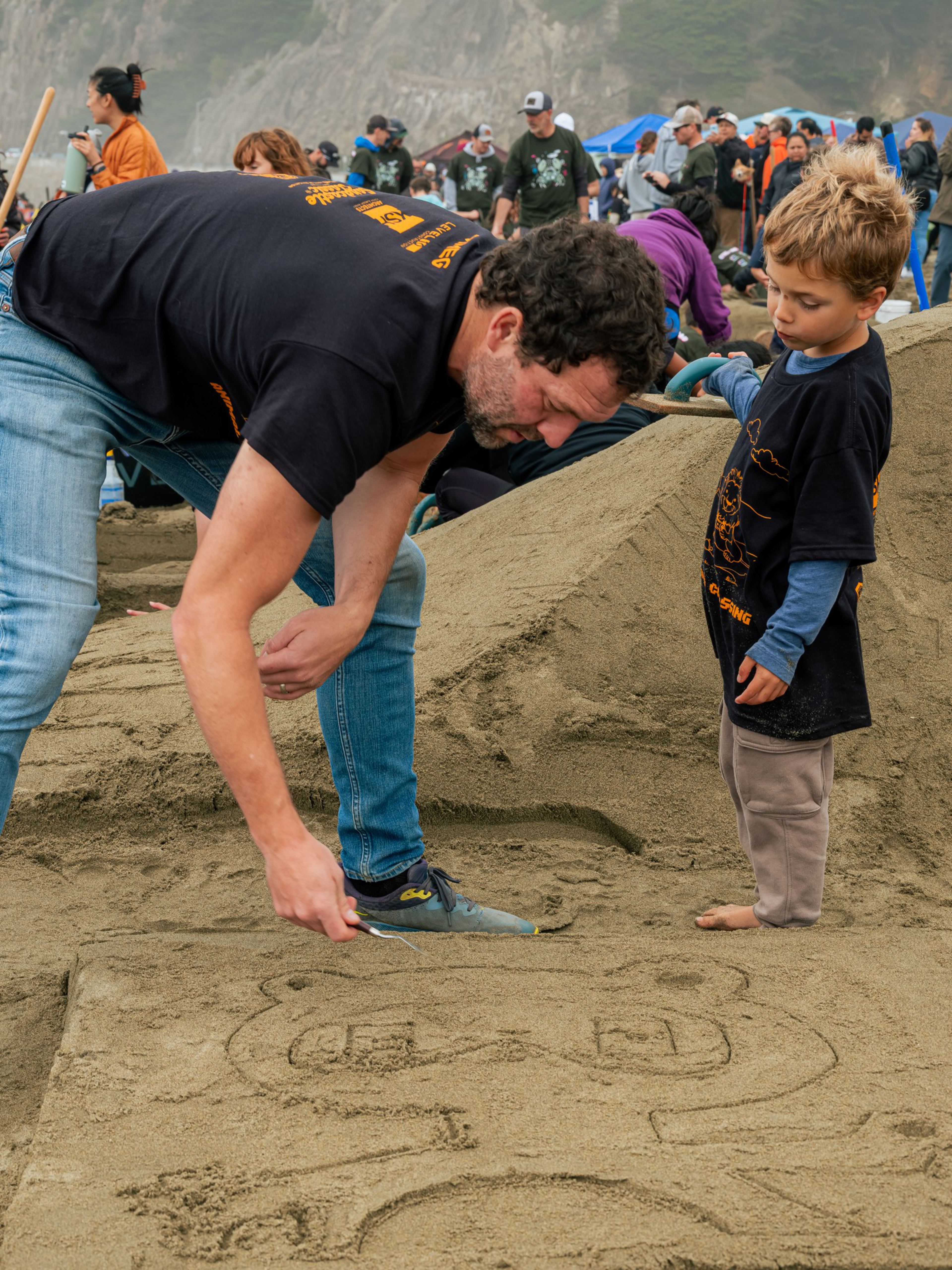 A man and a child wearing matching black shirts carve a bear face into the sand while a crowd gathers behind them.