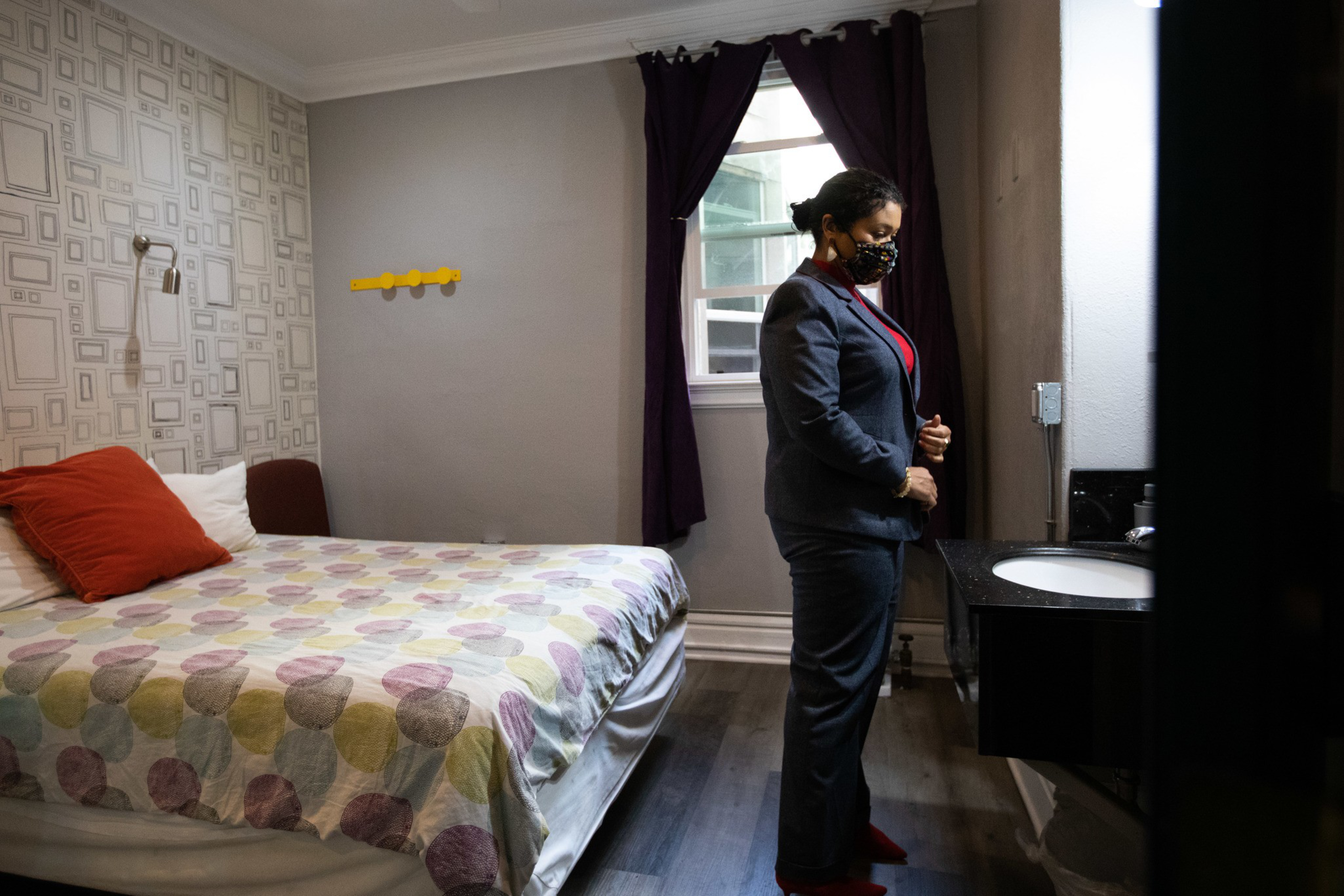 A woman in a suit and mask stands beside a bed with a colorful polka dot cover. The room has a small sink, purple curtains, and decorative wallpaper.