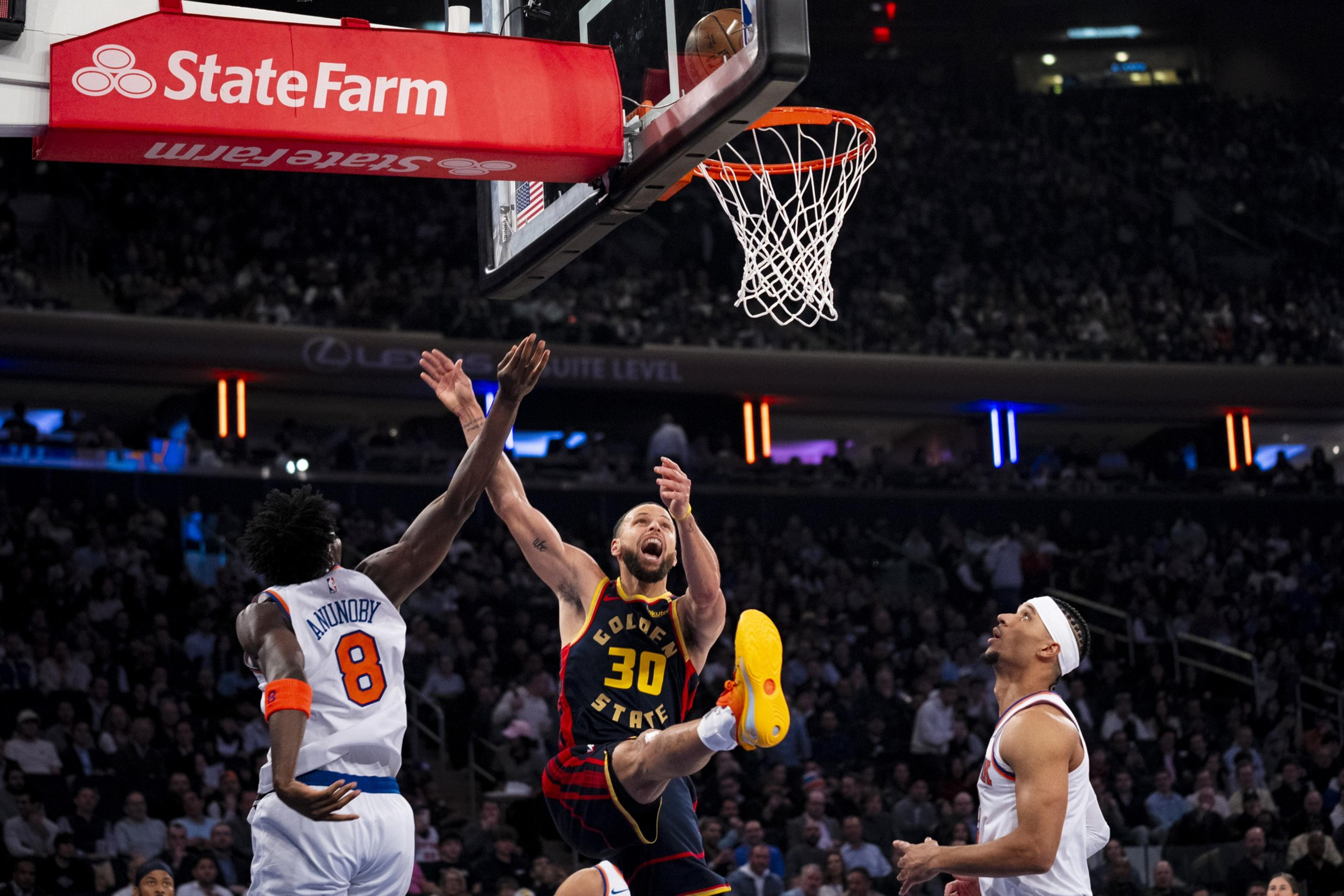 A basketball player in a dark jersey leaps towards the hoop, ball in hand, as opponents in white jerseys watch, under a bright red State Farm banner.