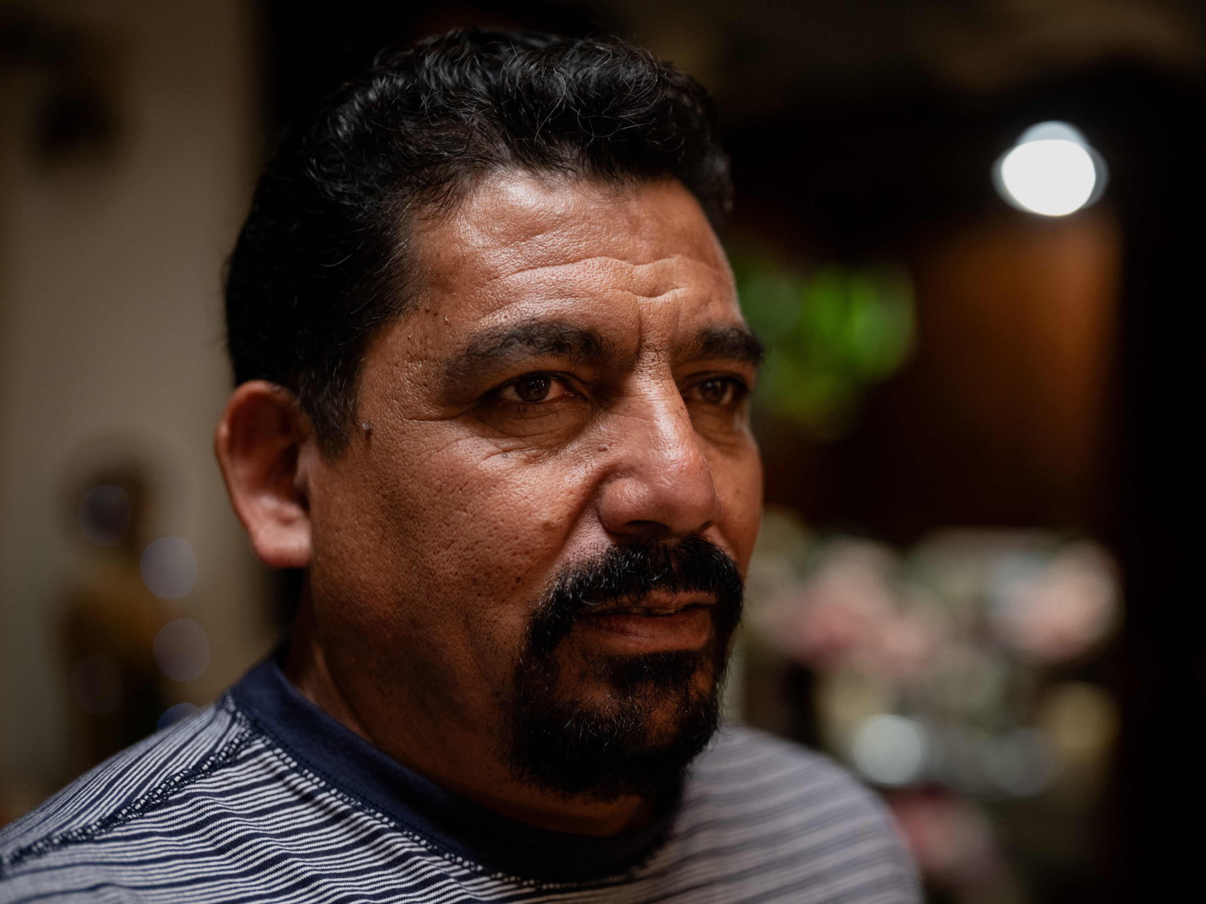 A middle-aged man with curly black hair, a thick mustache, and beard wears a striped shirt, looking thoughtfully to the side with a blurred warm background.