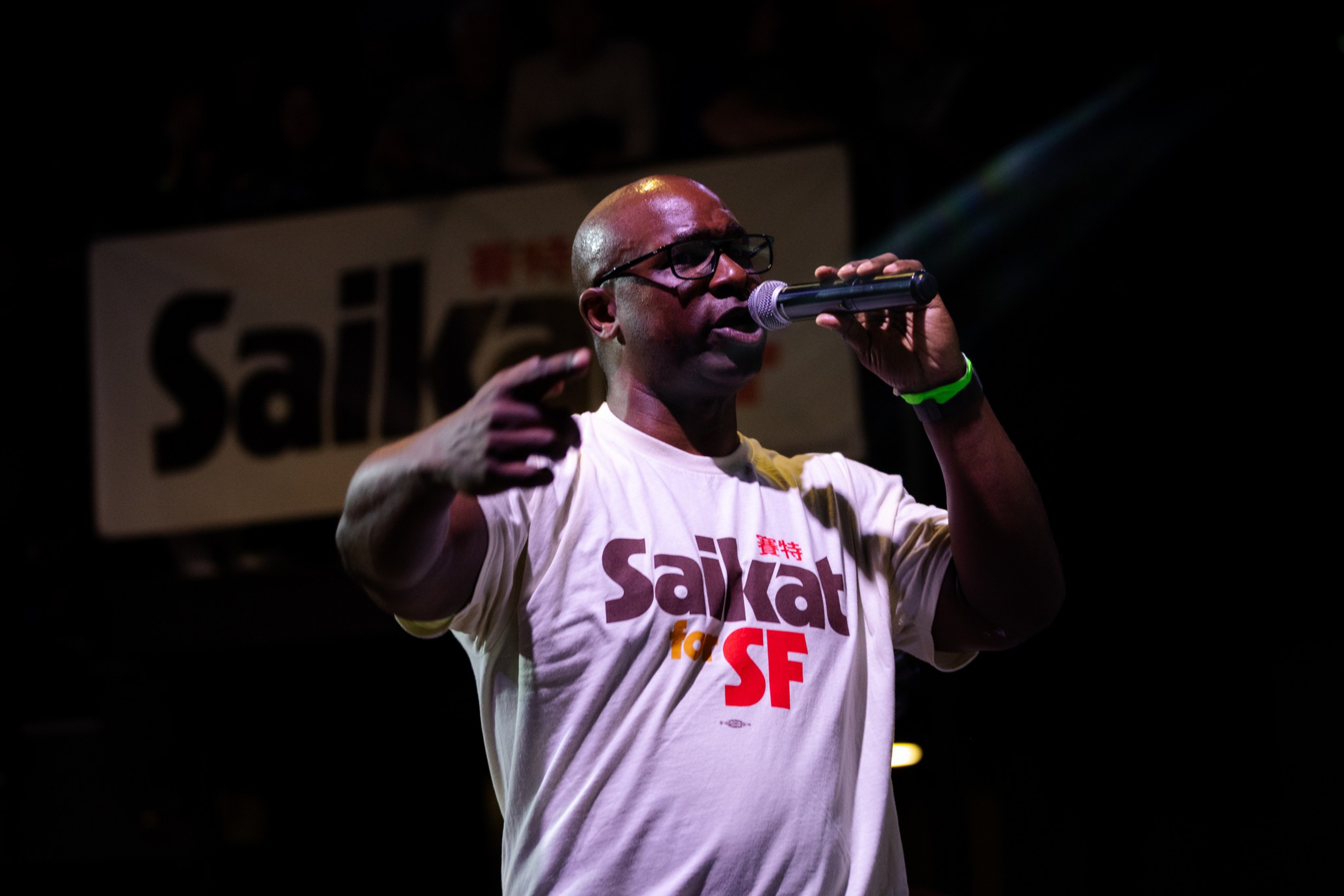 A man wearing glasses and a “Saikat for SF” shirt speaks into a microphone on stage with a matching banner in the background.