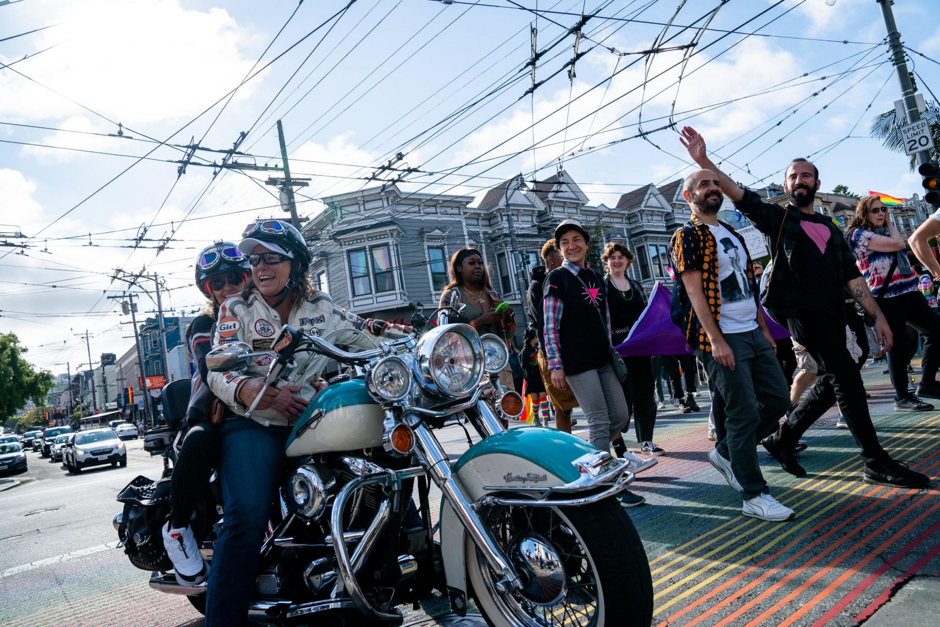 Two people wearing helmets ride a classic motorcycle near a crosswalk with rainbow stripes while diverse pedestrians walk beside them, one waving.