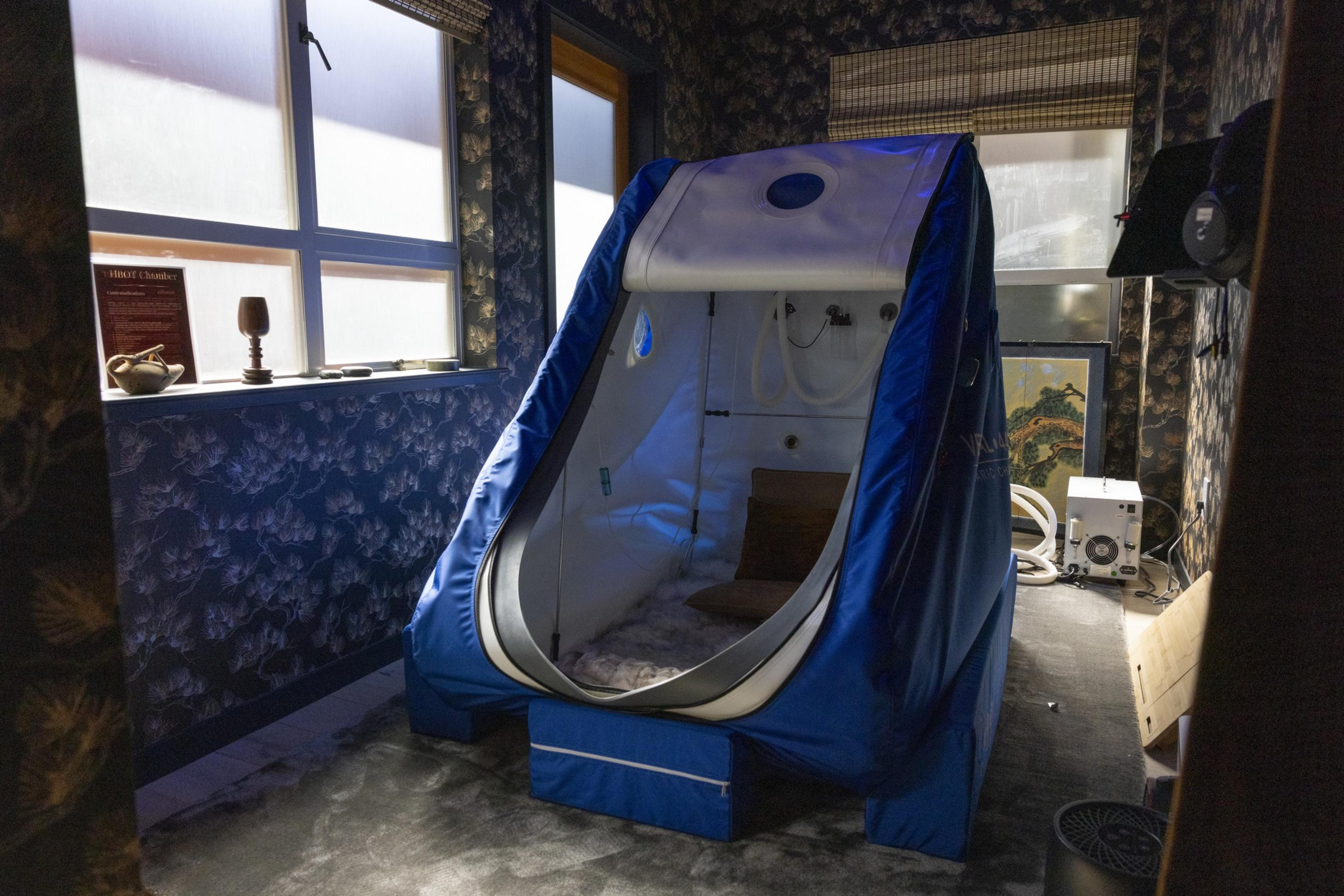The room contains a blue hyperbaric oxygen chamber next to a window with floral wallpaper and various small objects on the sill. The chamber is partially open, revealing its interior.