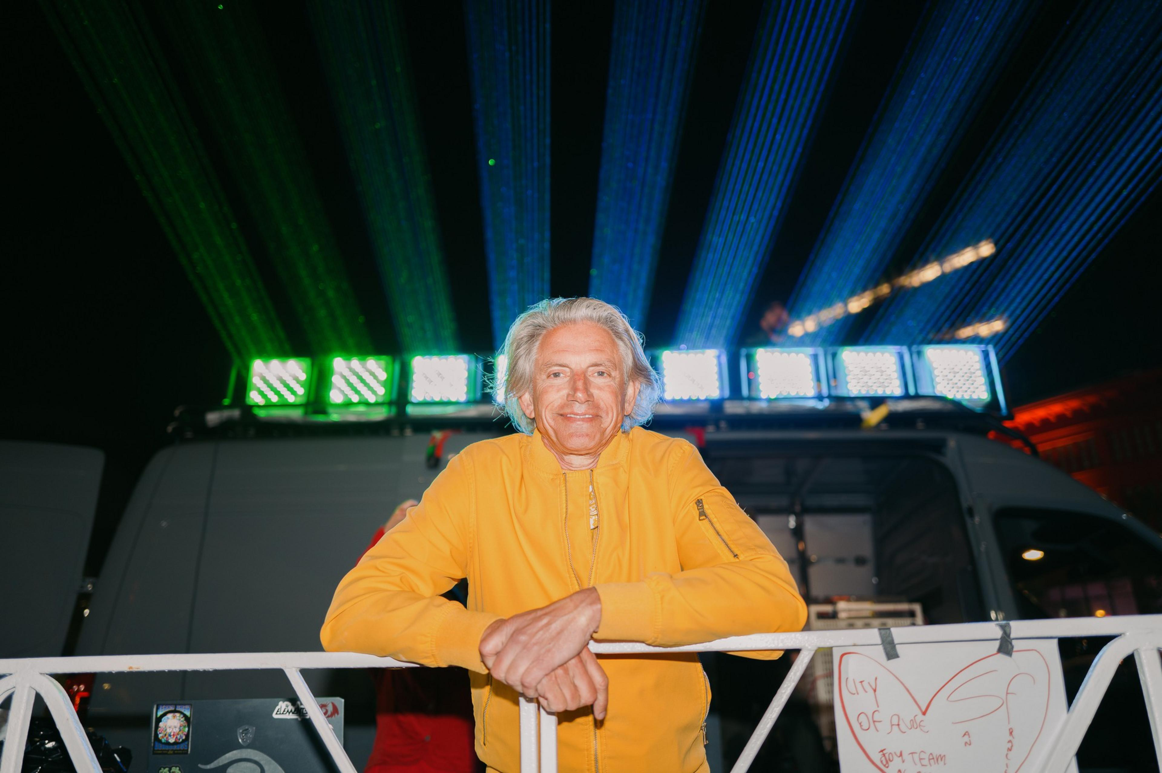 A person in a yellow jacket stands smiling with arms resting on a railing. Behind them, colorful laser lights create an eye-catching display in the dark.
