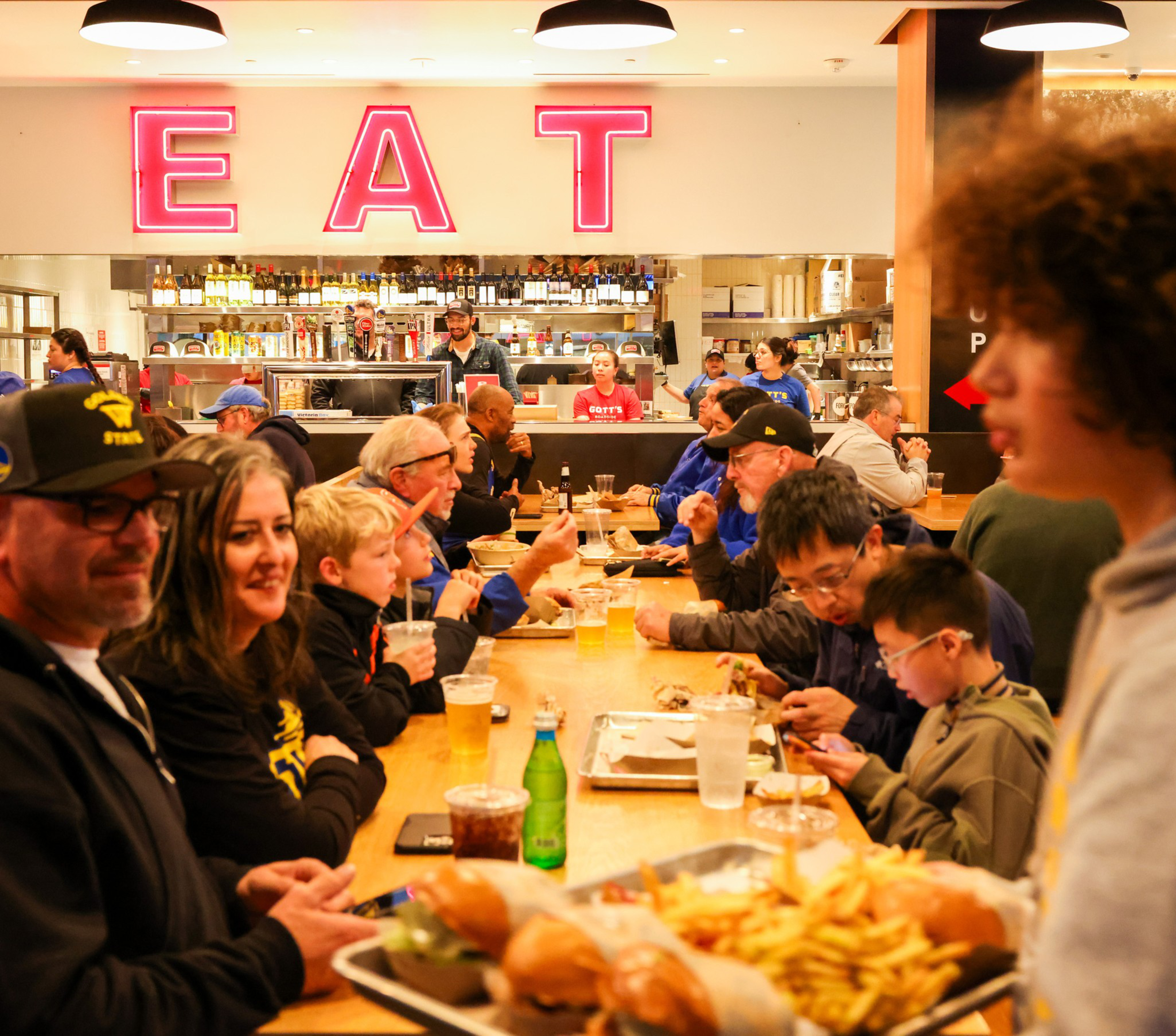 A group of people are dining at a long table in a lively restaurant. A large neon sign reads &quot;EAT&quot; above a busy kitchen, and trays of food and drinks are visible.