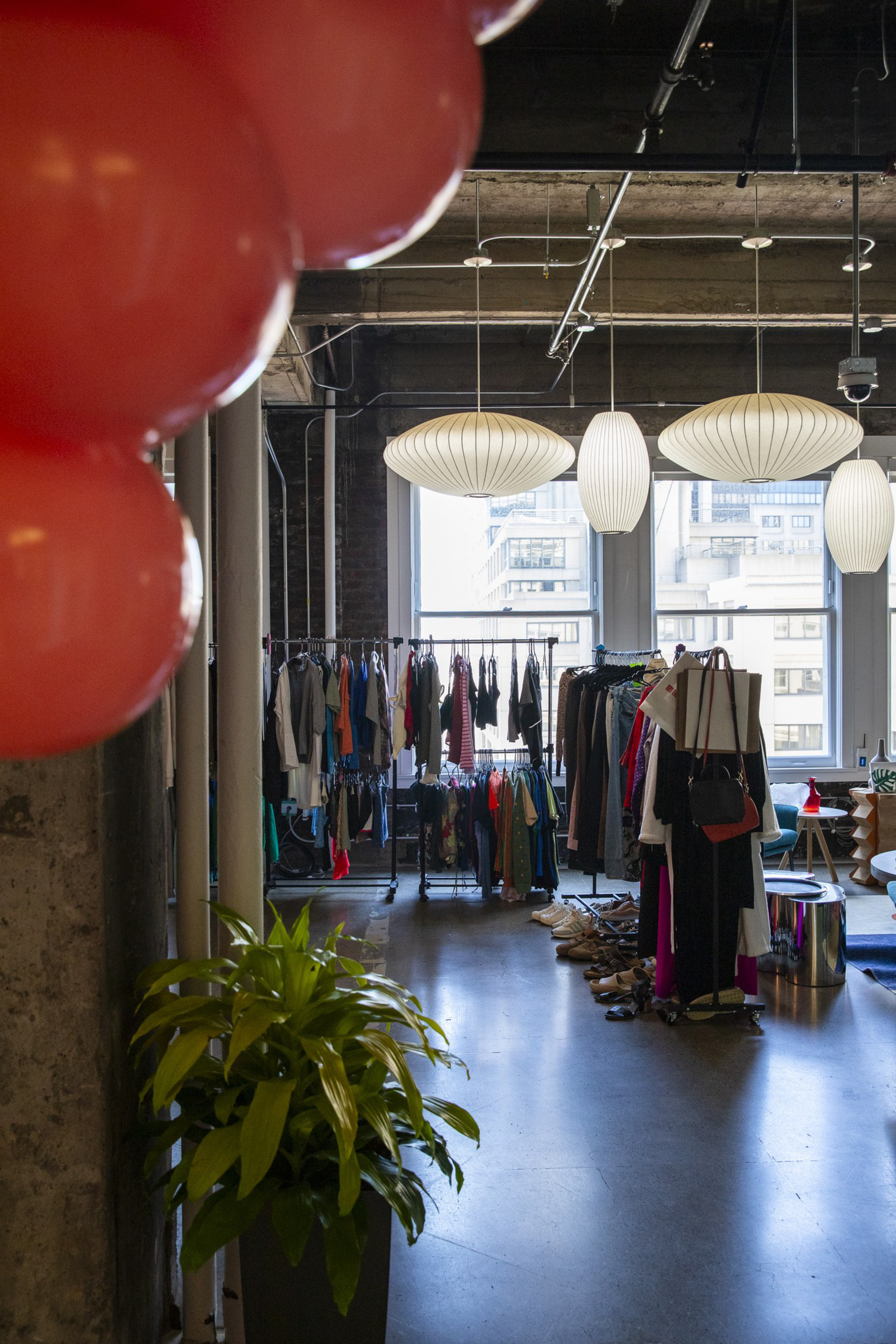 An industrial-style room with clothing racks, shoes on the floor, large windows, hanging white lamps, a leafy green plant, and red balloons in the foreground.