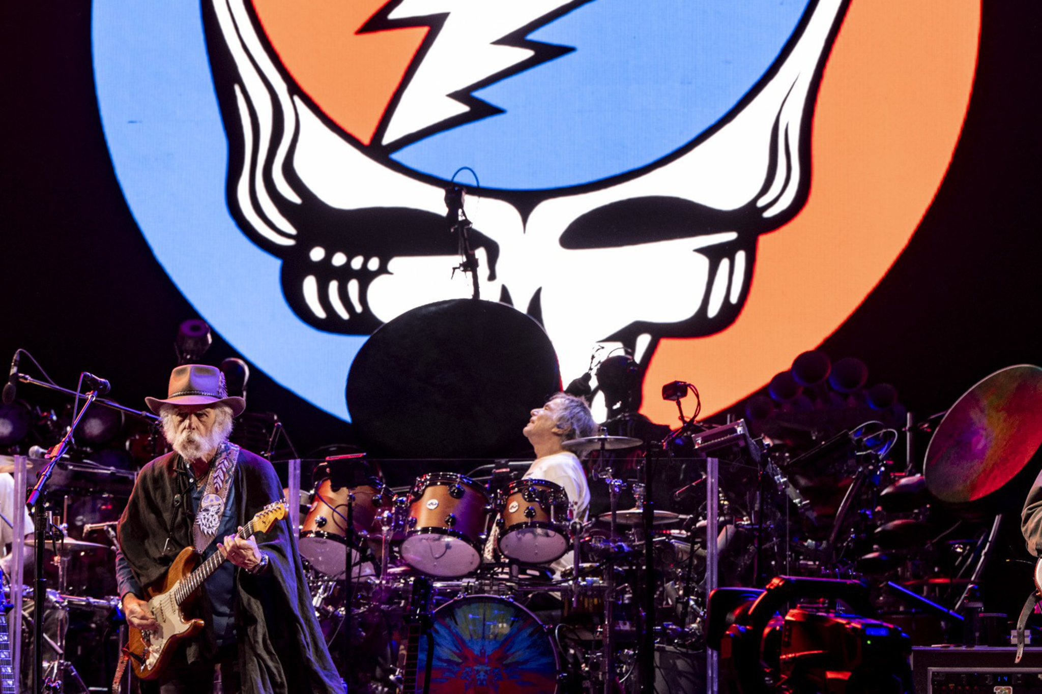 A musician with a guitar and a drummer perform on stage in front of a large, colorful skull logo split in blue and red with a white lightning bolt.