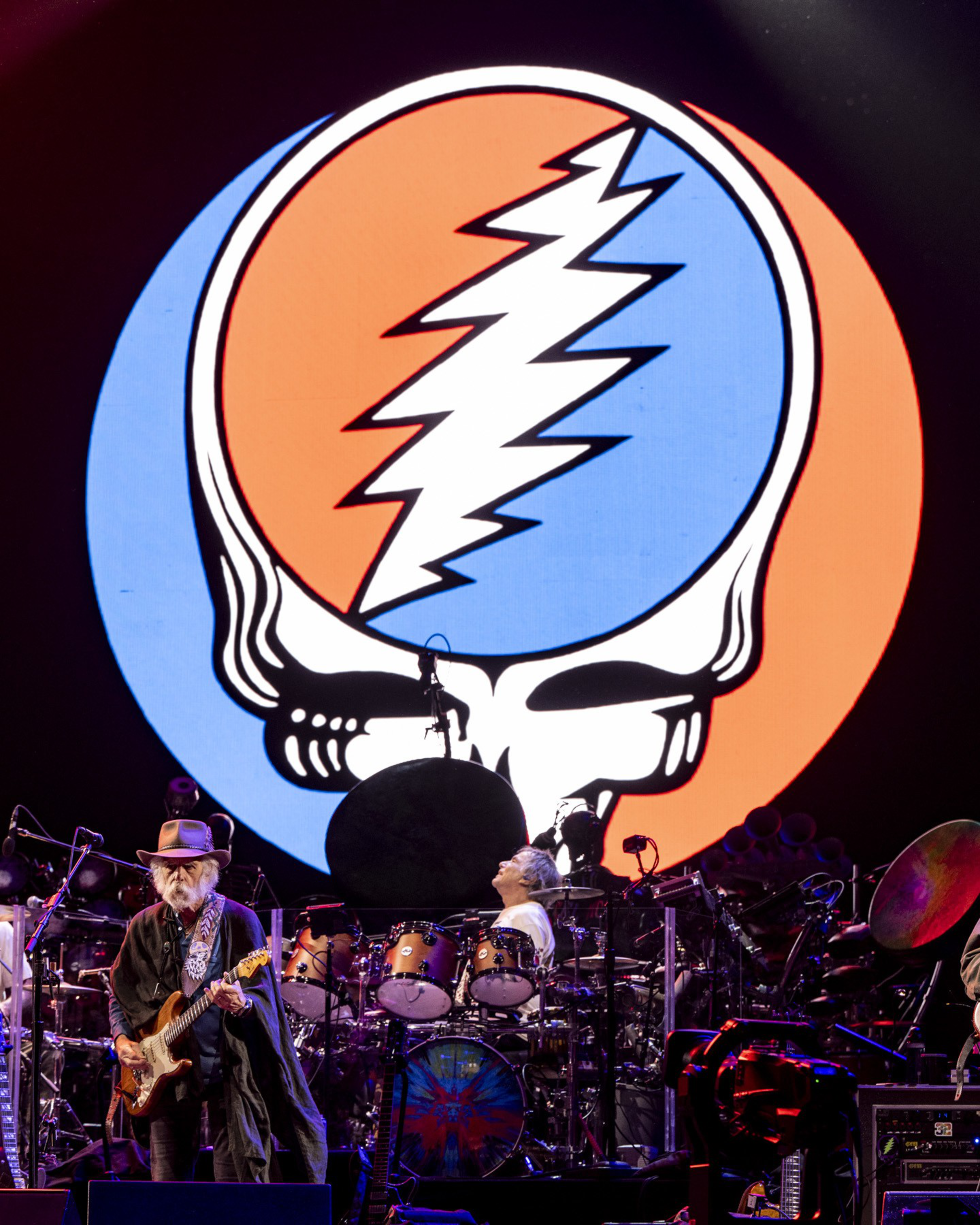 A musician with a guitar and a drummer perform on stage in front of a large, colorful skull logo split in blue and red with a white lightning bolt.