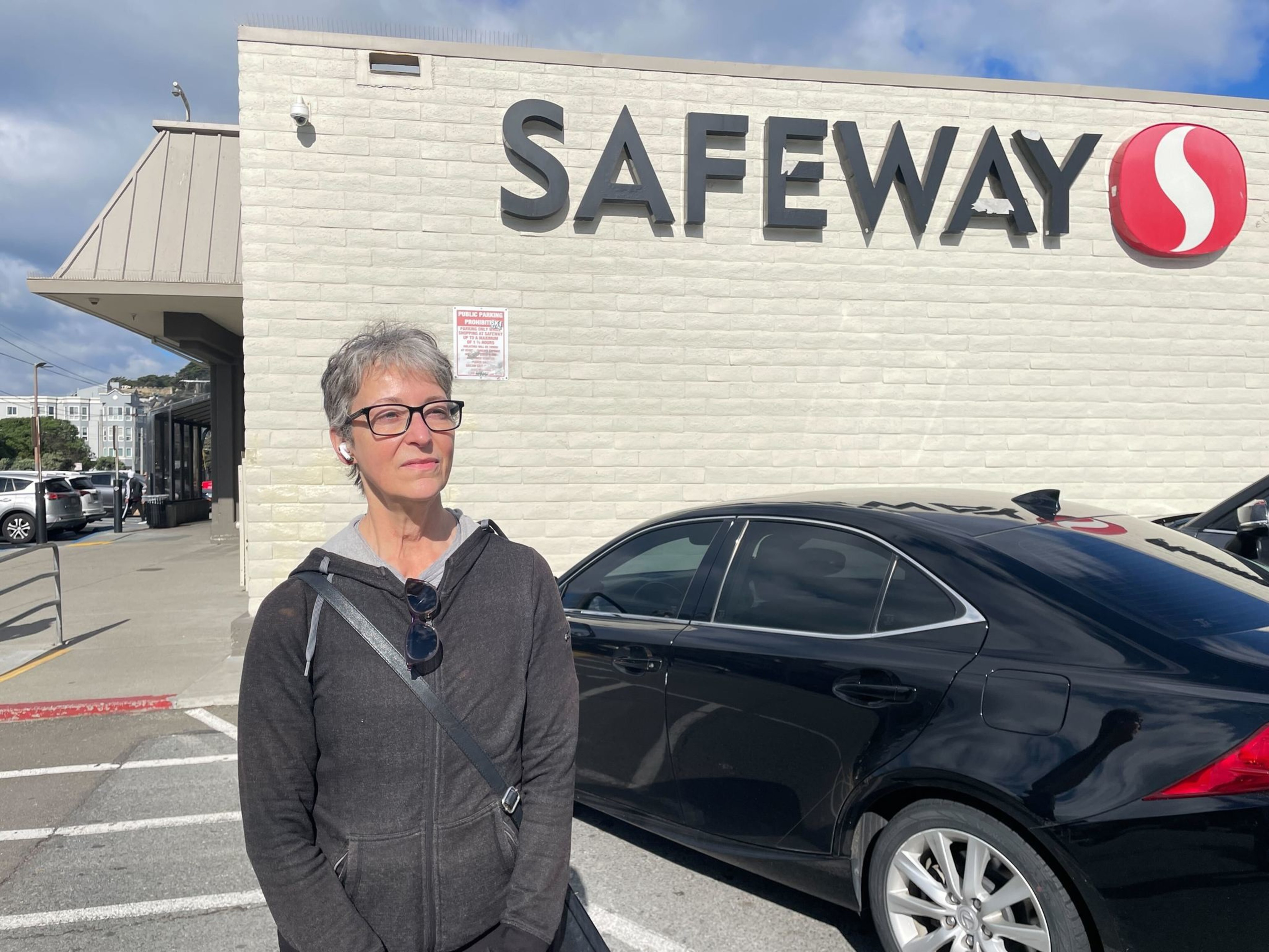 A woman wearing glasses and a dark hoodie stands near a black car in front of a Safeway supermarket on a sunny day.