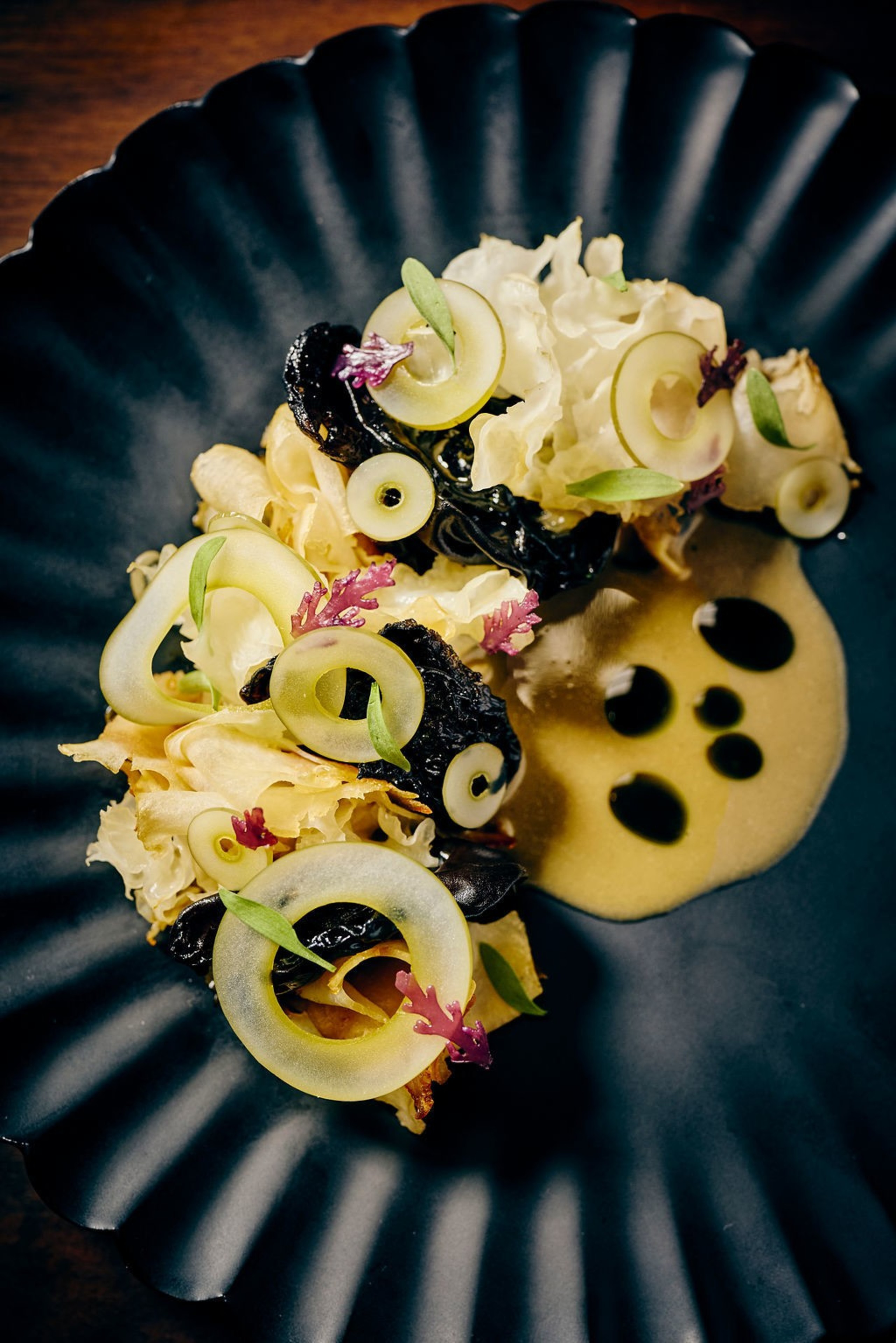 Delicately arranged on a black plate, this dish features thin slices of grapes and decorative greens atop a bed of white and dark leafy vegetables in sauce.